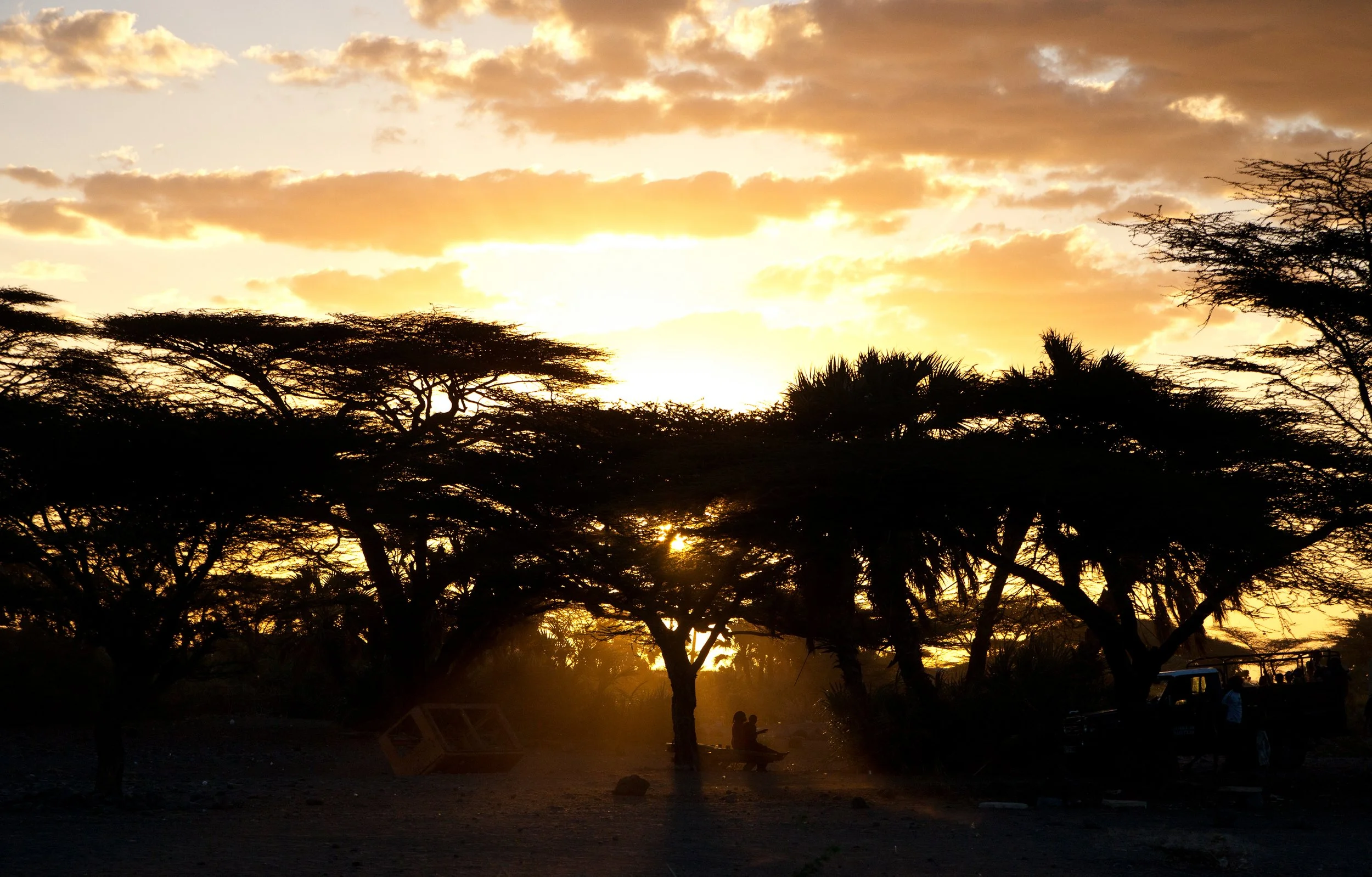 Sonnenuntergang in der afrikanischen Savanne mit Baumkronen, zwei Personen sitzen auf einer Parkbank, ein Fahrzeug und eine Person stehen im Hintergrund.