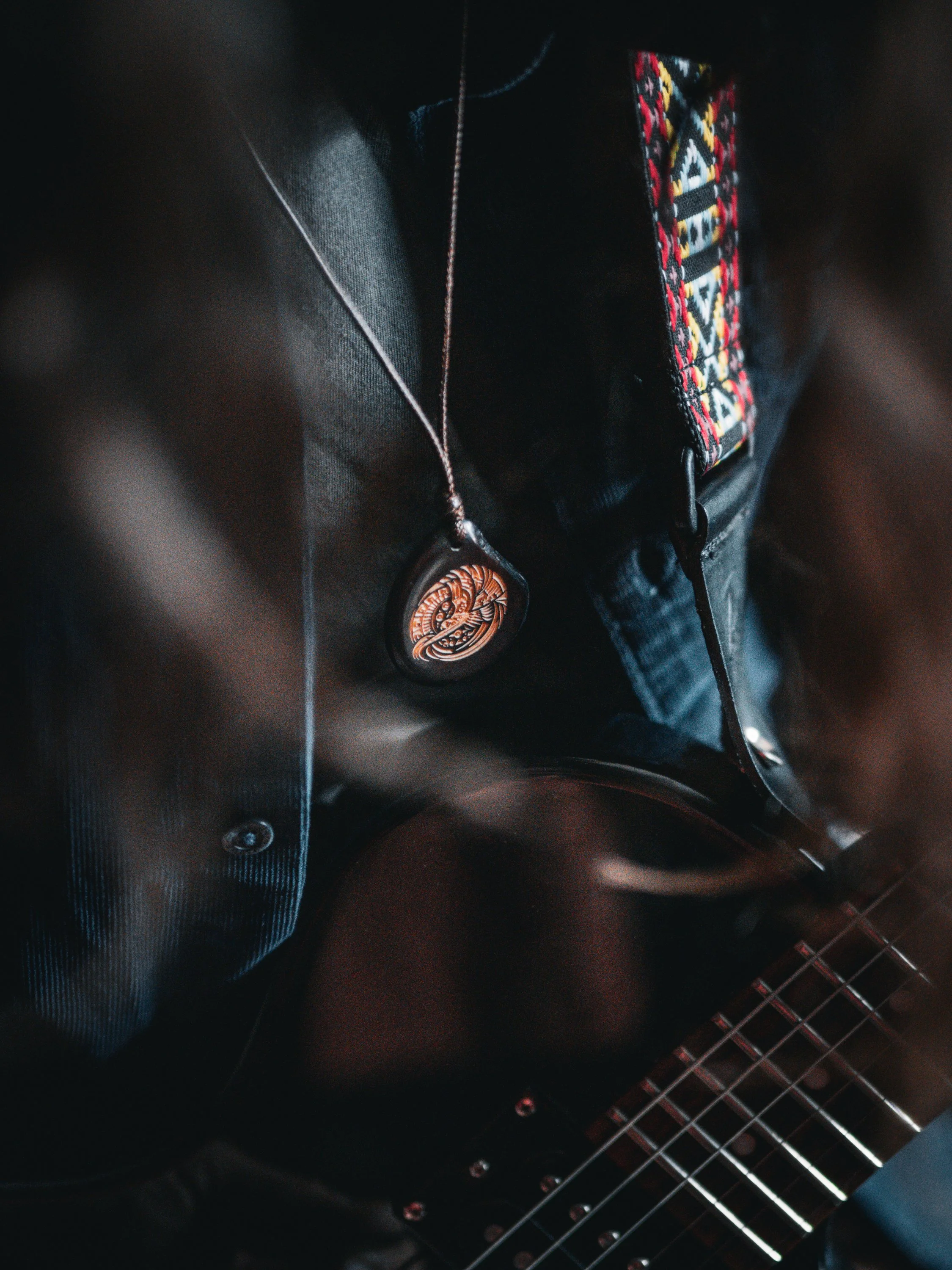 Nahaufnahme eines Menschen mit einer E-Gitarre, getragen mit einem schwarzen Lederjacke, einem silbernen Halskettenanhänger und einer farbenfrohen, gestickten Tasche auf der Brust