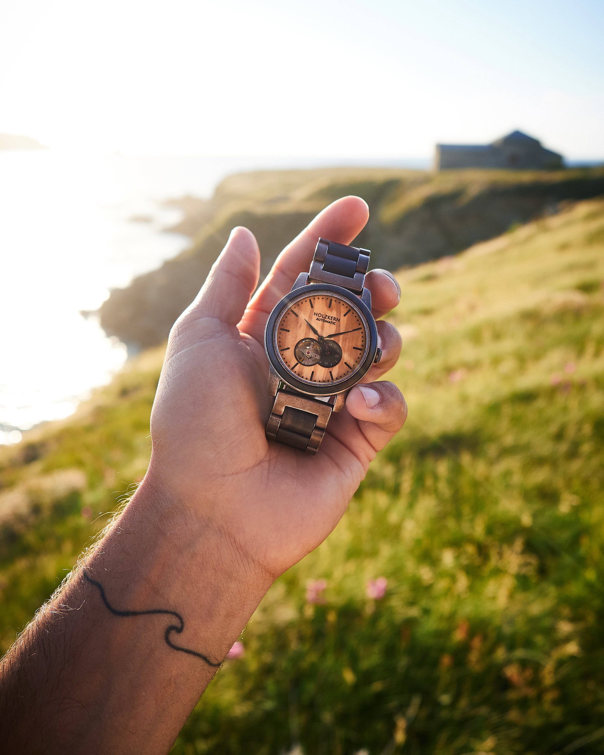 Hand hält eine Armbanduhr mit braunem Zifferblatt vor einer Küstenlandschaft bei Sonnenuntergang.