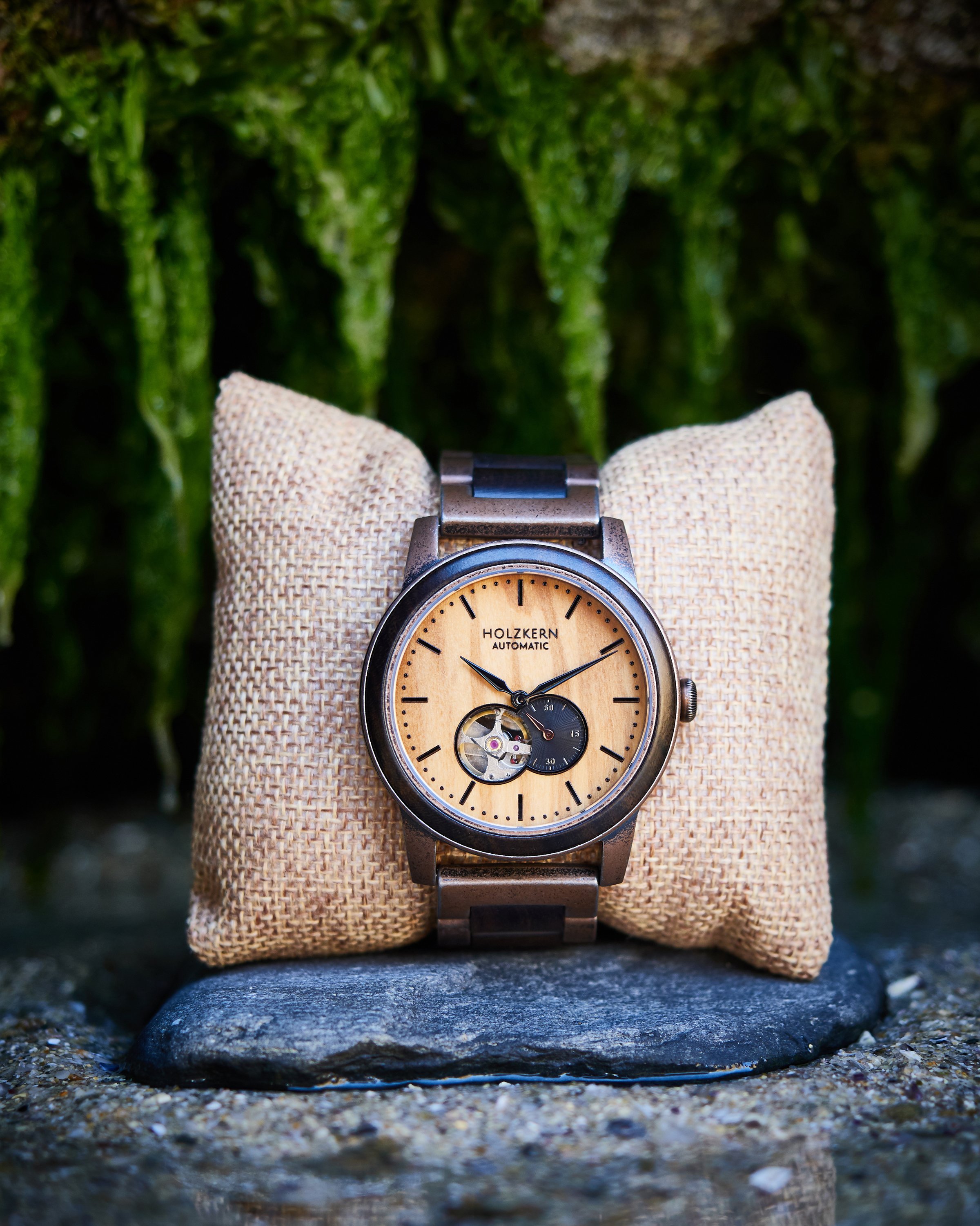 Armbanduhr mit holzfarbenem Zifferblatt auf einem Stoffkissen, im Freien, mit grünen Moos im Hintergrund.