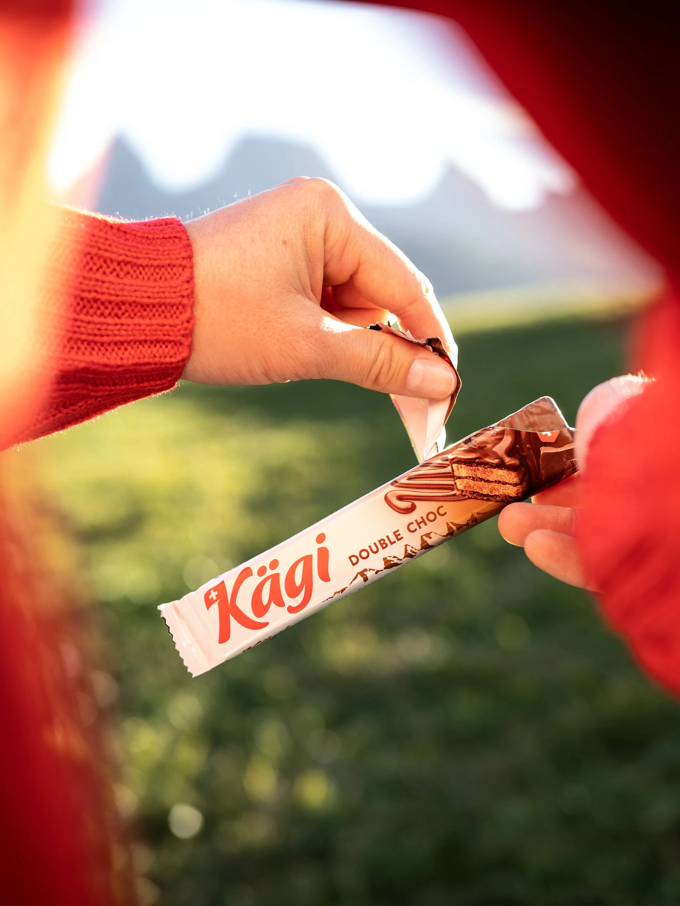 Eine Person in einem roten Pullover hält ein Kagi Double Choc Schokoriegel in der Hand, gesehen durch eine kleine Öffnung zwischen roten Stoffen, mit einer grünen Wiese und Bergen im Hintergrund.