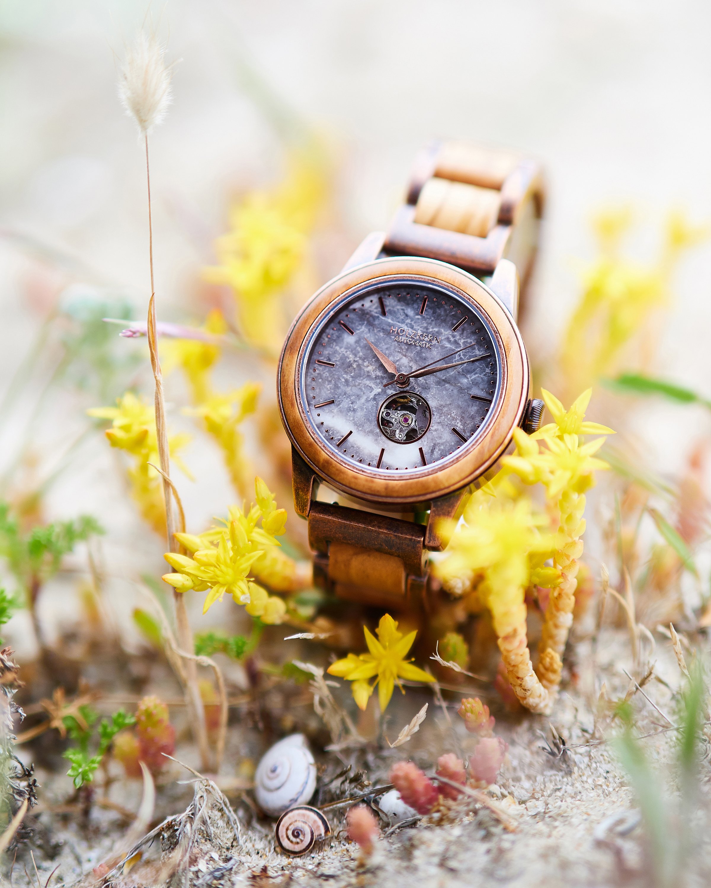 Eine Armbanduhr mit Holzgehäuse und Armband, die auf Sand mit gelben Blumen und Muscheln liegt.