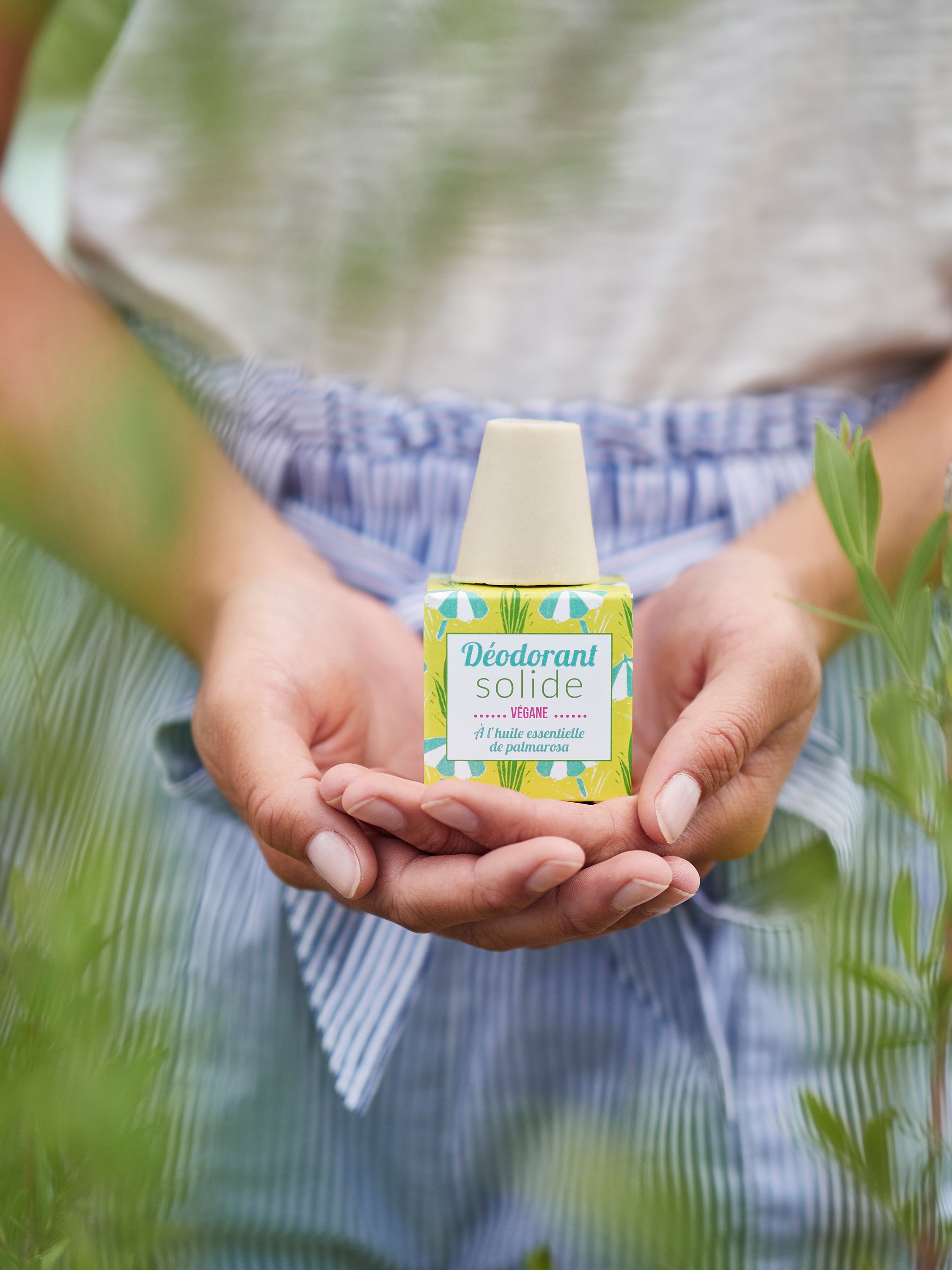 Hand hält eine Flasche vegane Deodorantcreme mit grünen und blauen Verpackungsmotiv inmitten von Pflanzen.