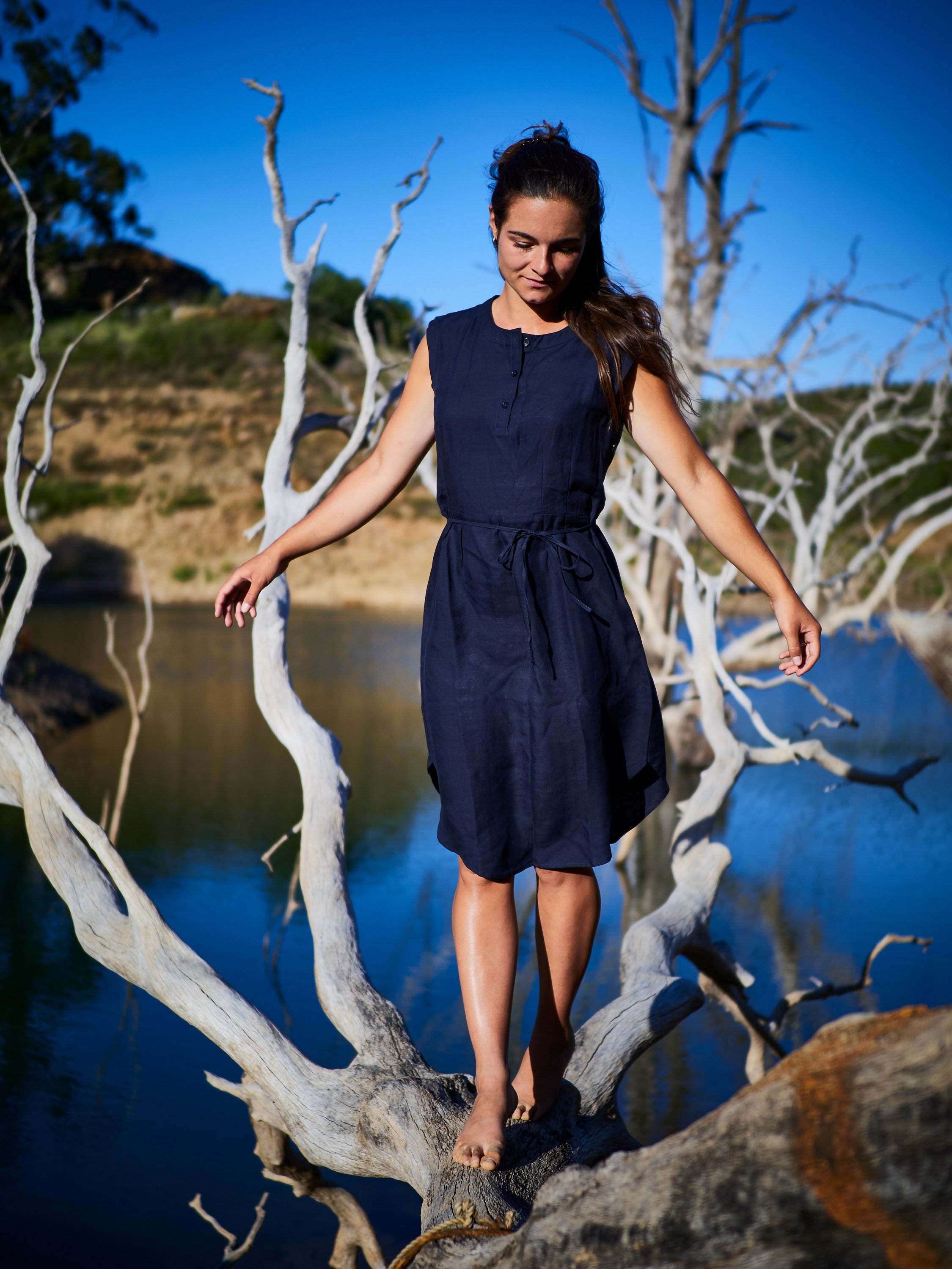 Eine junge Frau in einem dunkelblauen Kleid balanciert barfuß auf einem Ast eines toten Baumes am Wasser mit blauem Himmel im Hintergrund.