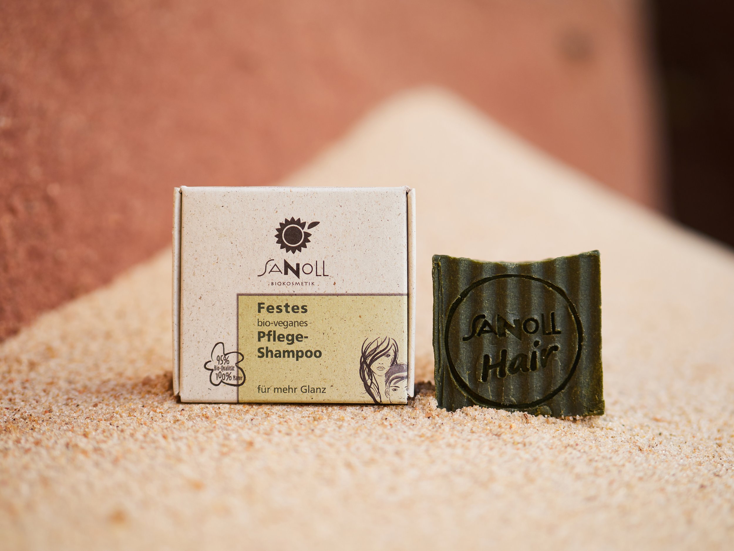 Bio veganes Festes Pflege-Shampoo in einer Pappschachtel und einem grünen Seifenstück mit eingedrucktem Schriftzug, beide auf Sand liegend, im Hintergrund Sanddünen.