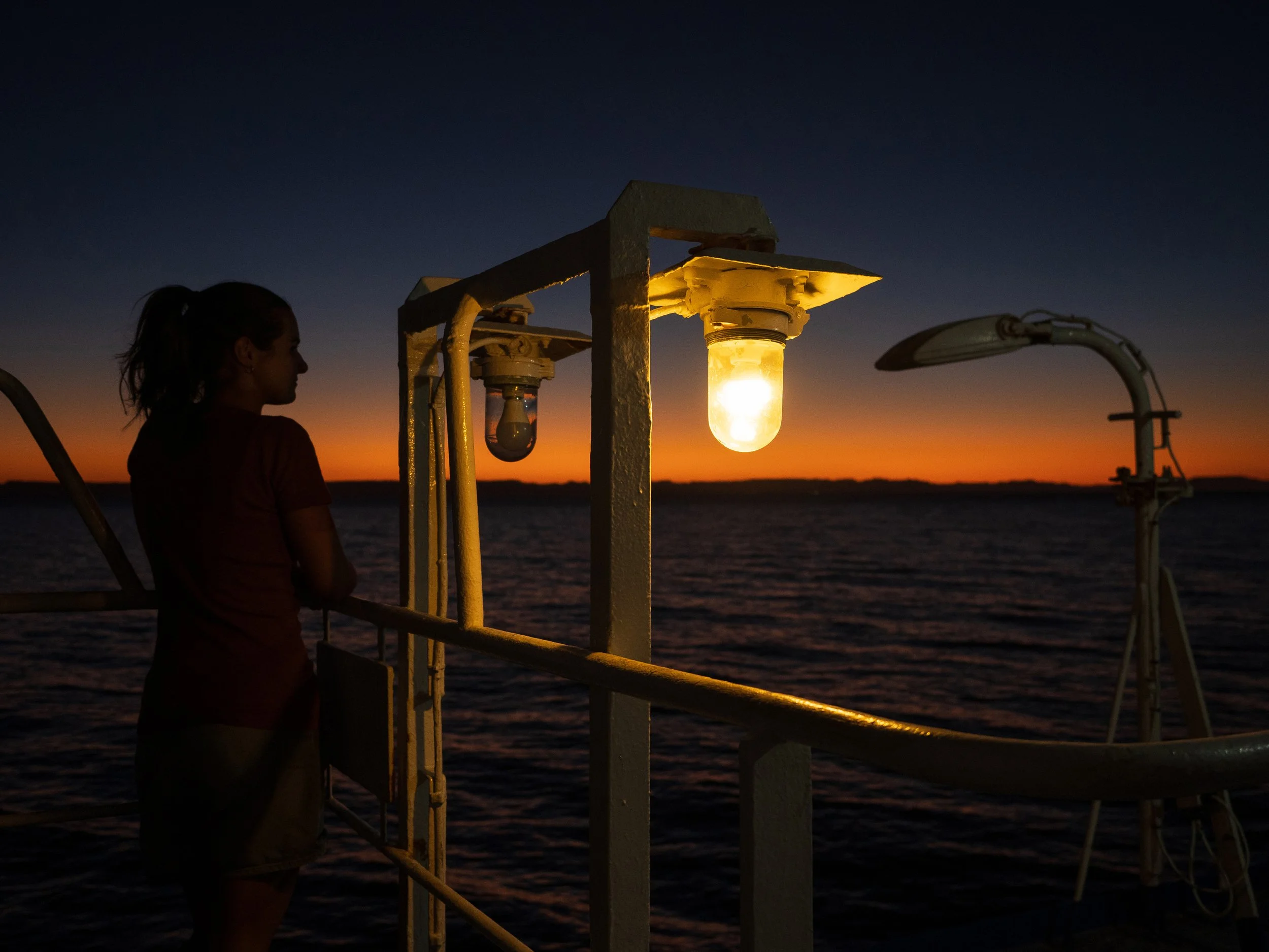 Frau auf einem Boot bei Sonnenuntergang, beleuchtete Lampen auf dem Deck, ruhiges Wasser im Hintergrund.