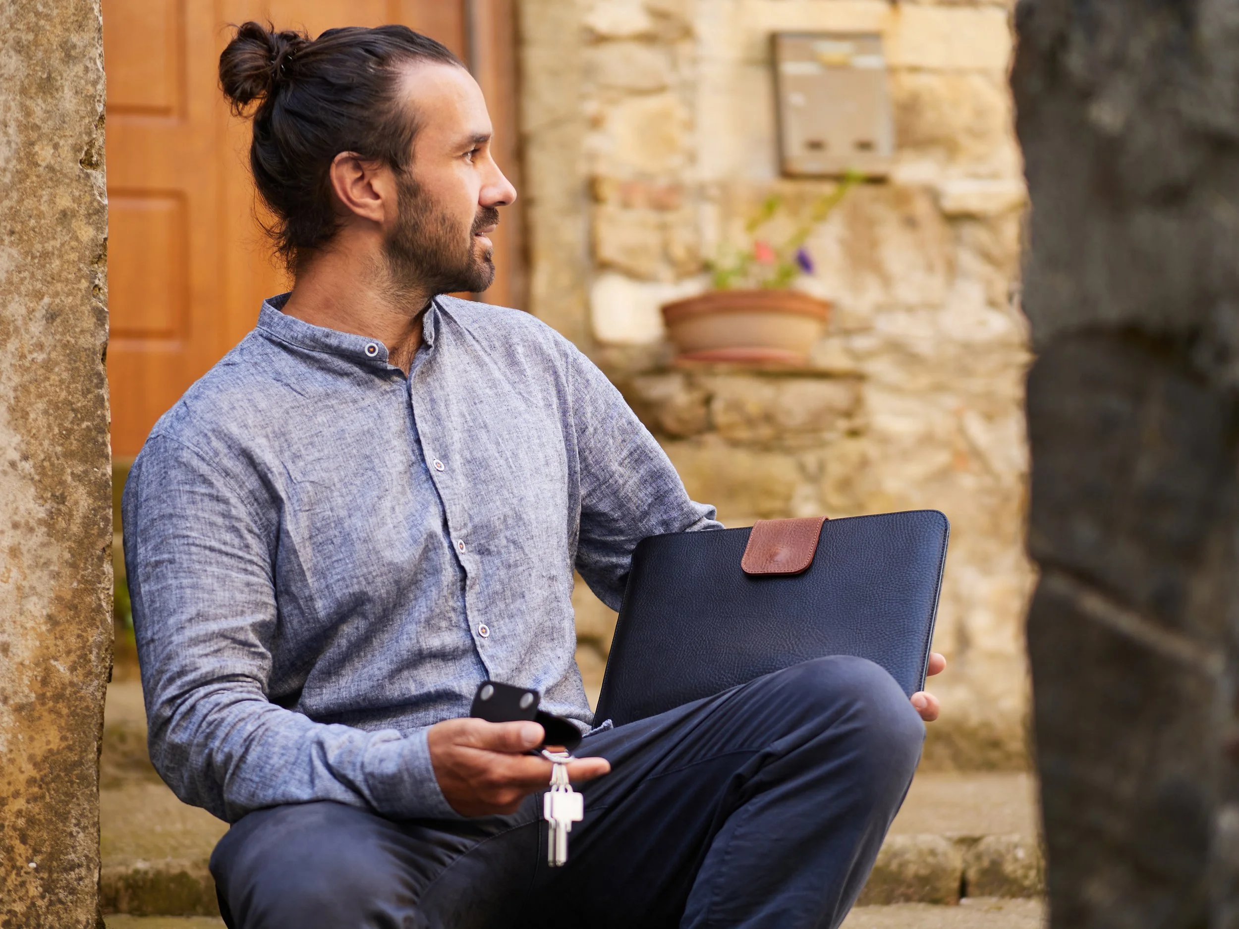 Ein Mann sitzt draußen an einer Steinmauer, hält einen Schlüsselbund und ein Smartphone, trägt eine graue Hemdjacke und hat einen Laptop auf dem Schoß.