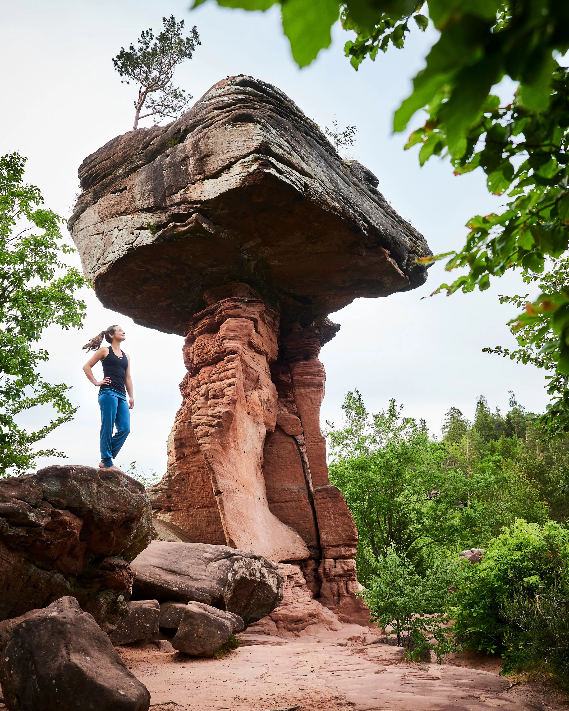 Eine junge Frau steht auf einem Felsen vor einer großen, beeindruckenden roten Felsformation in einer grünen, bewaldeten Umgebung.