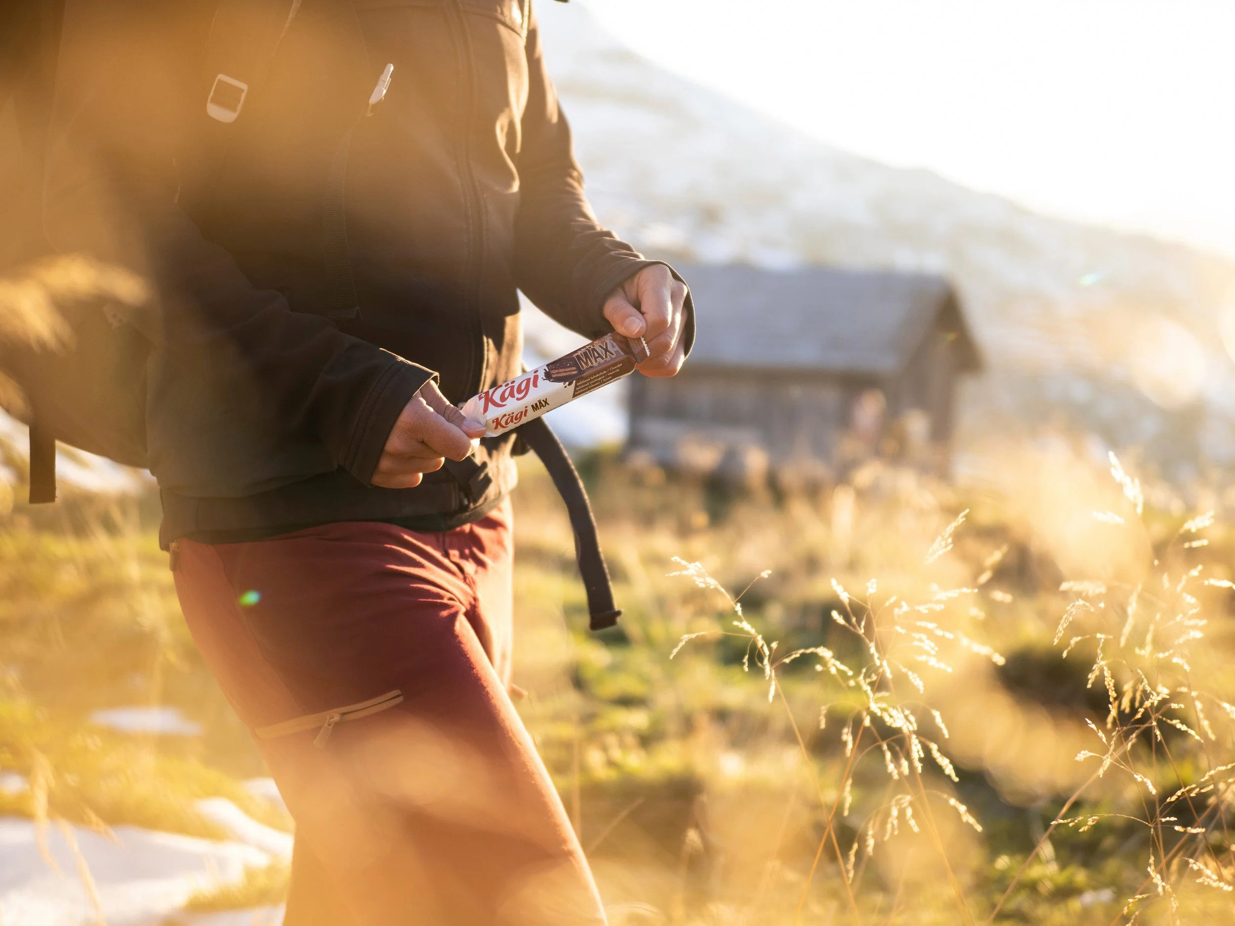 Person beim Wandern im Gelände, hält eine Schokolade namens "Kägi MAX" in der Hand, mit Sonnenlicht und einer Hütte im Hintergrund.