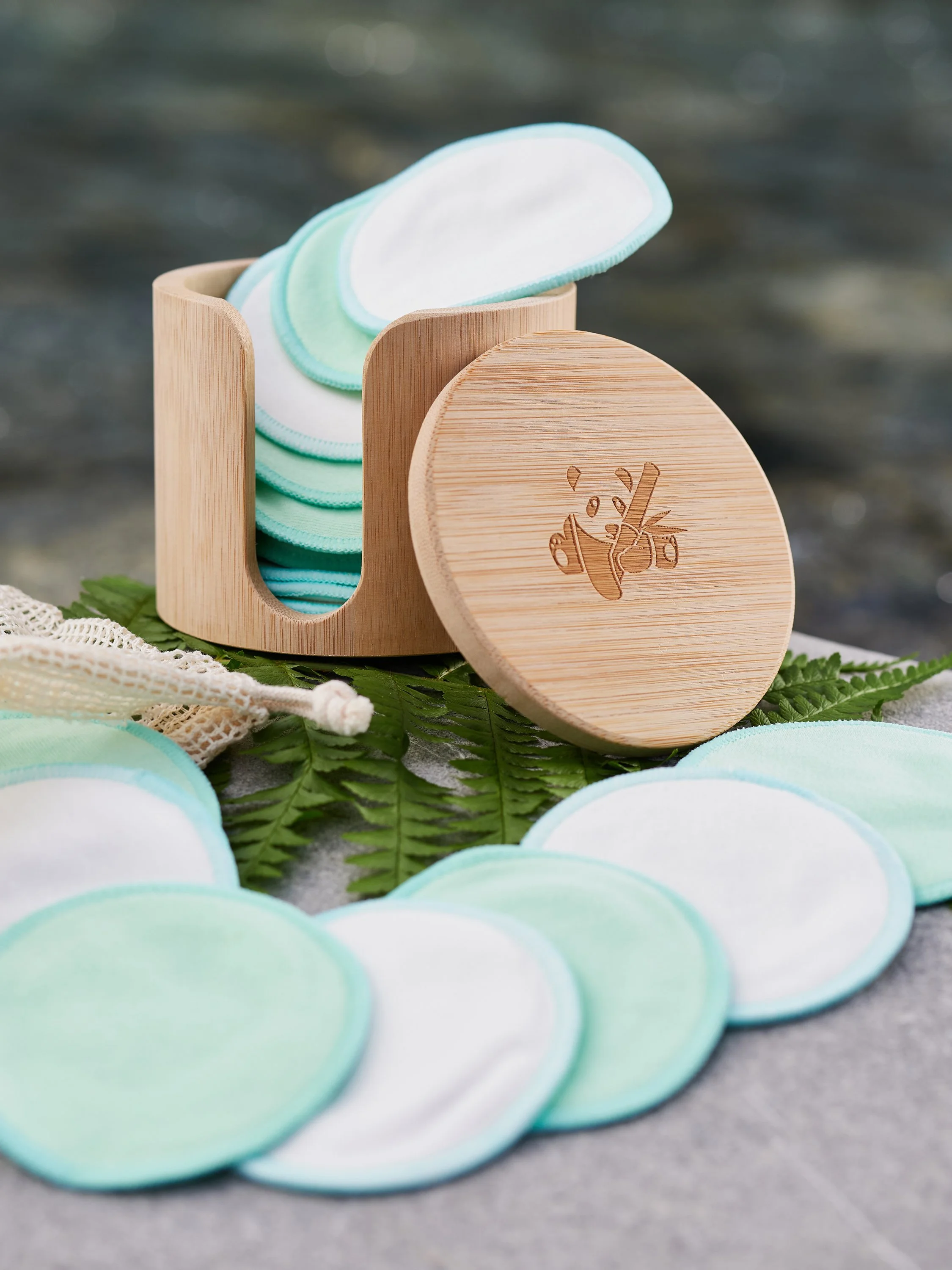 Runde, waschbare Stoffuntersetzer in Pastellfarben (blau, grün, weiß) liegen auf einem Felsen, daneben eine Holzdose mit weiteren Untersetzern, die aus Bambusholz mit eingraviertem Logo bestehen, samt einem kleinen Stoffbeutel.