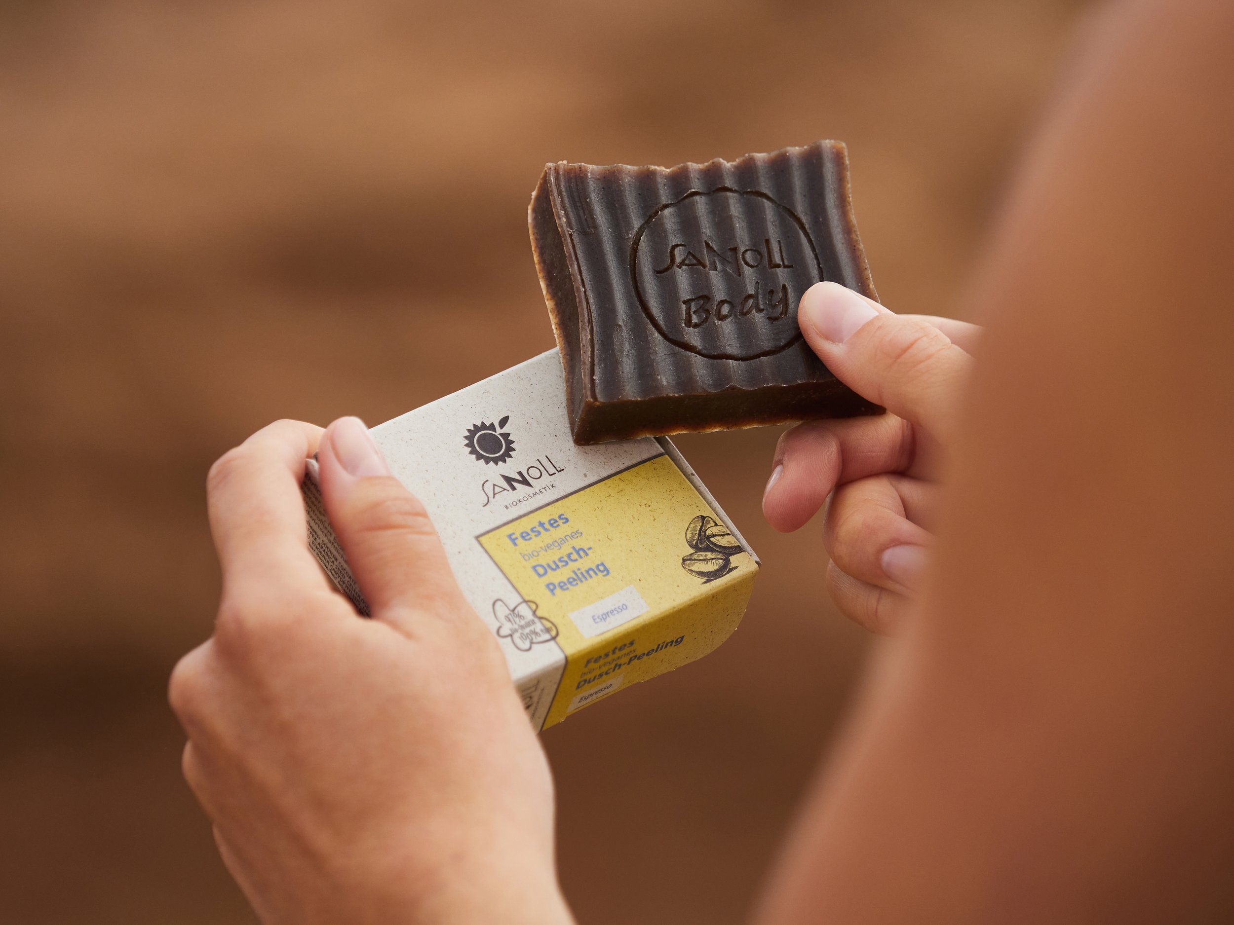 Nahaufnahme einer Person, die ein Stück dunkle Seife mit der Aufschrift 'Sanoll Body' hält, sowie eine Verpackung mit dem Label 'Sanoll' und der Beschreibung 'Festes Bio-veganes Dusch Peeling'.