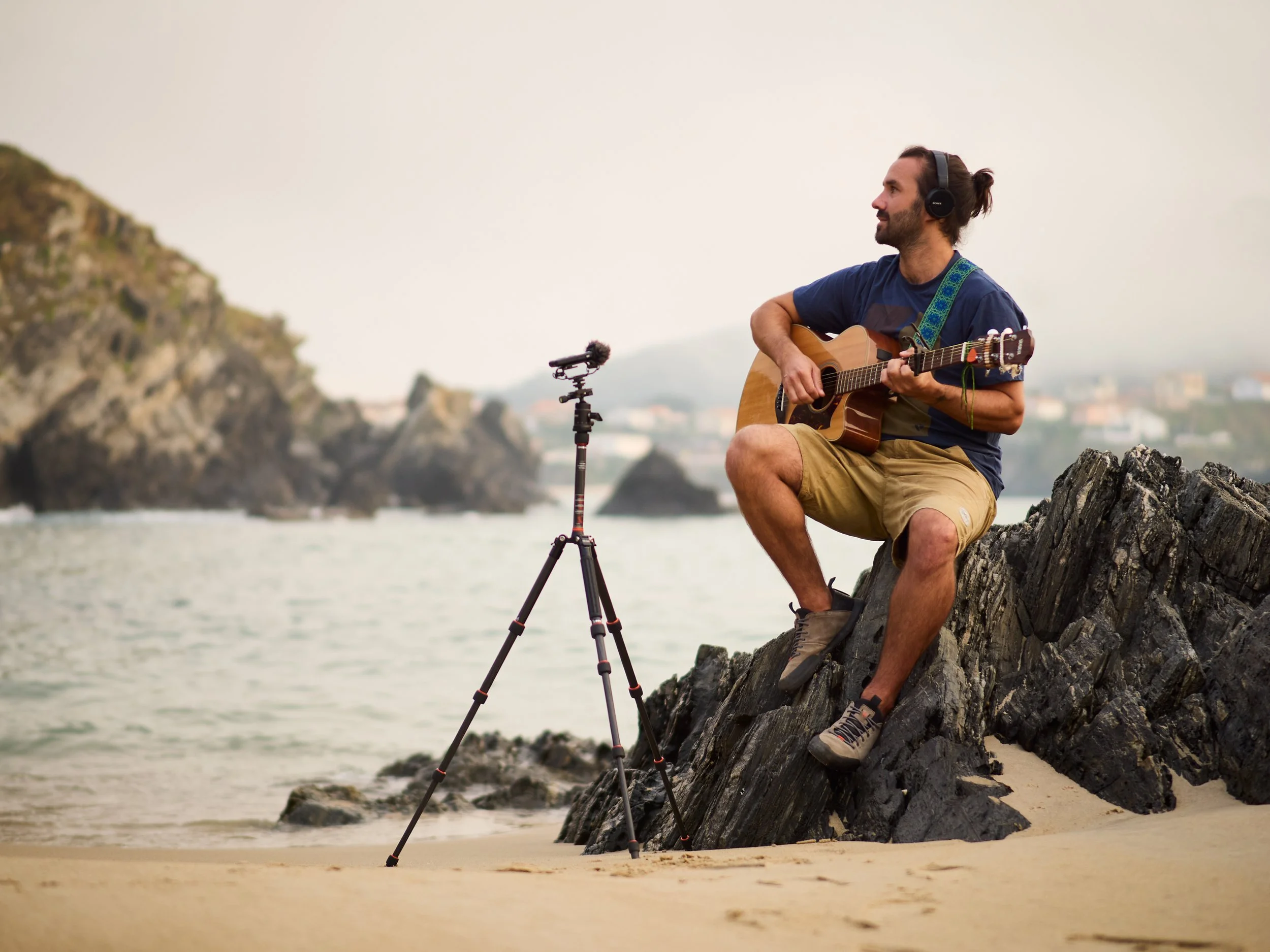 Mann spielt Gitarre am Strand, trägt Kopfhörer, sitzt auf Felsen, mit Kamera auf Stativ, im Hintergrund das Meer und Felsen an der Küste.