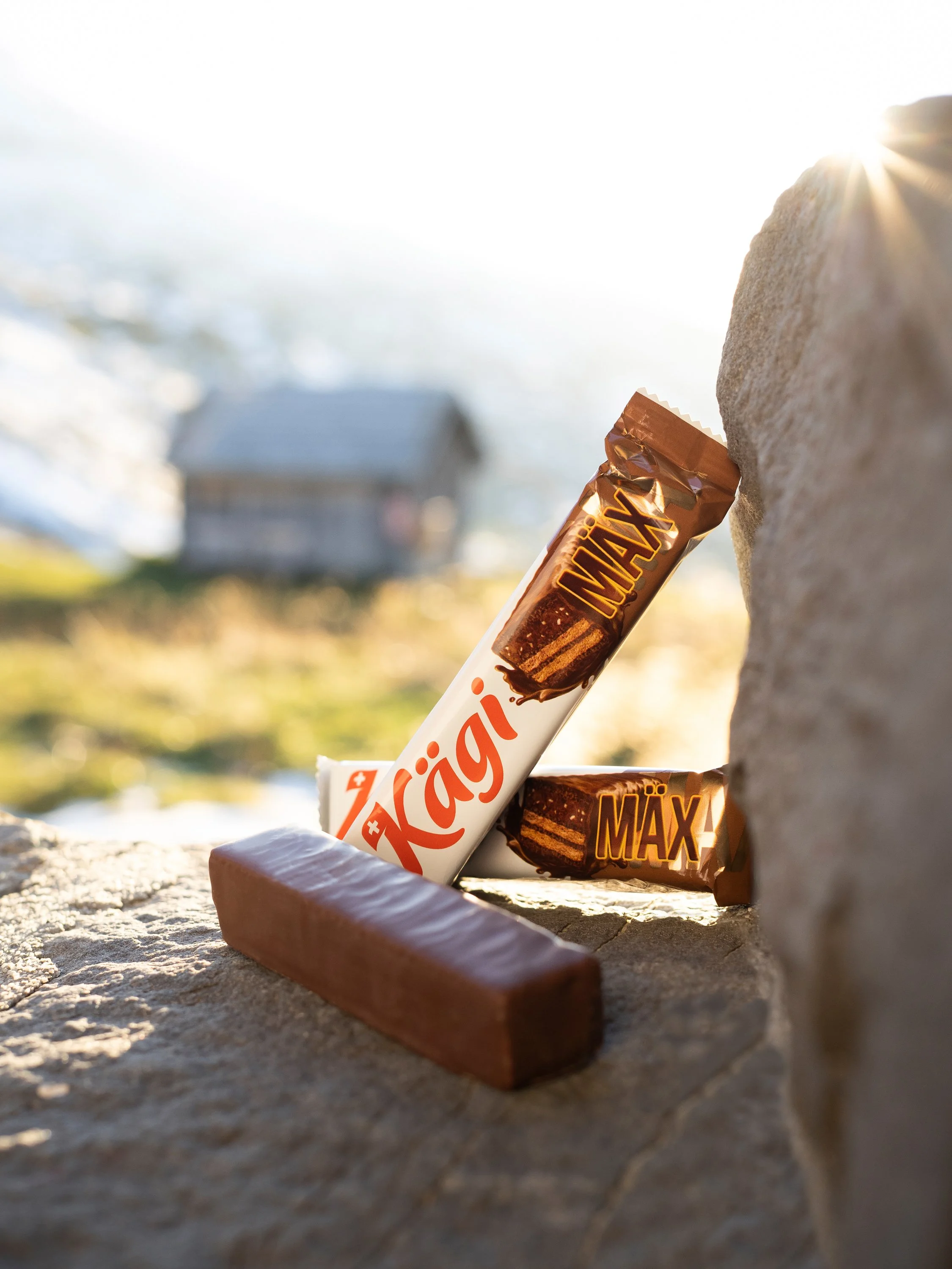 Zwei Riegel Schokolade, eine davon Käägi MAX, liegen auf einer Steinplatte im Freien bei Sonnenlicht; im Hintergrund ist eine Berglandschaft sichtbar.