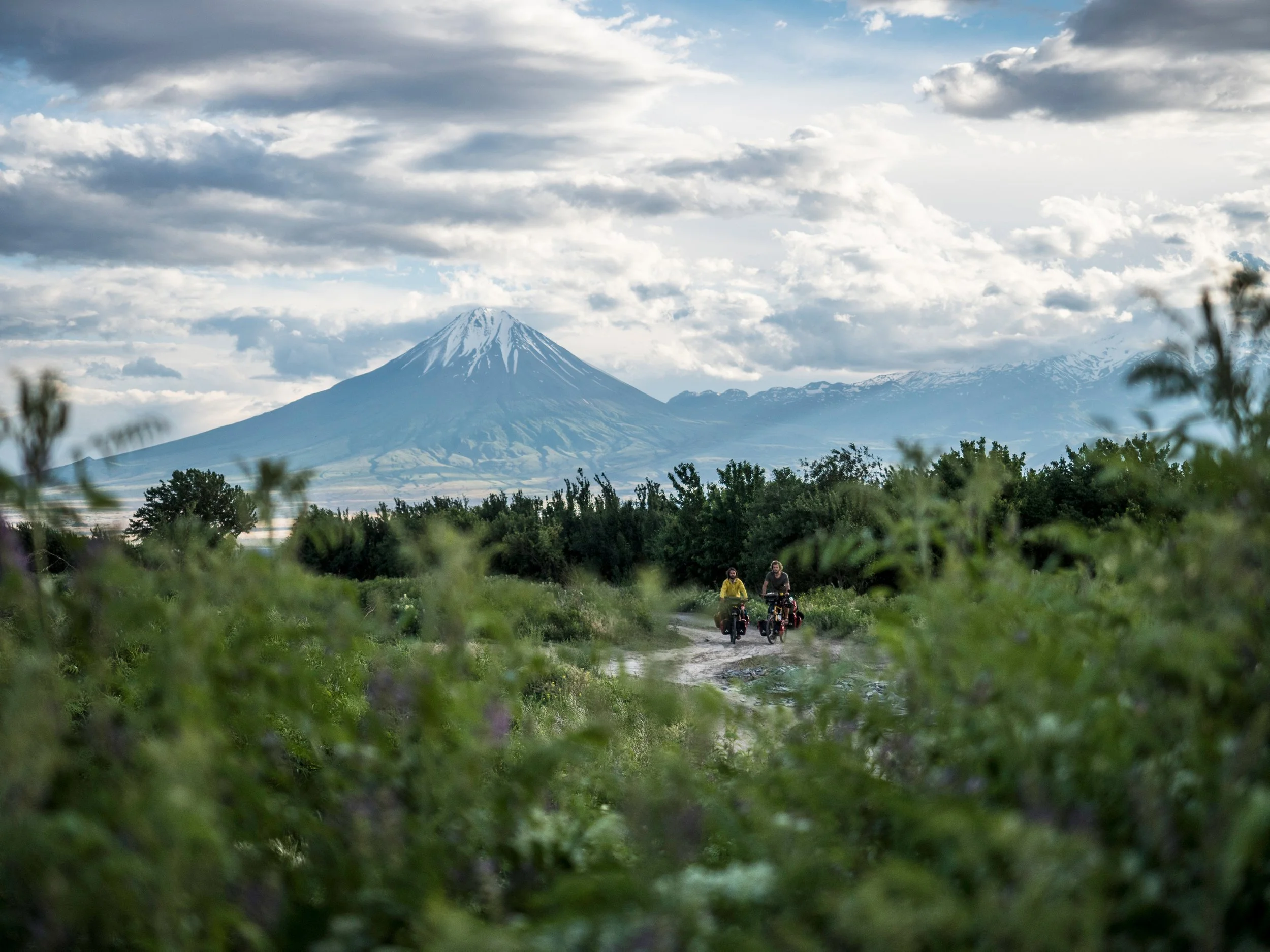 Zwei Radfahrer auf einem Weg mit Blick auf den schneebedeckten Mount Fuji im Hintergrund, umgeben von grünen Büschen und Bäumen unter einem bewölkten Himmel.
