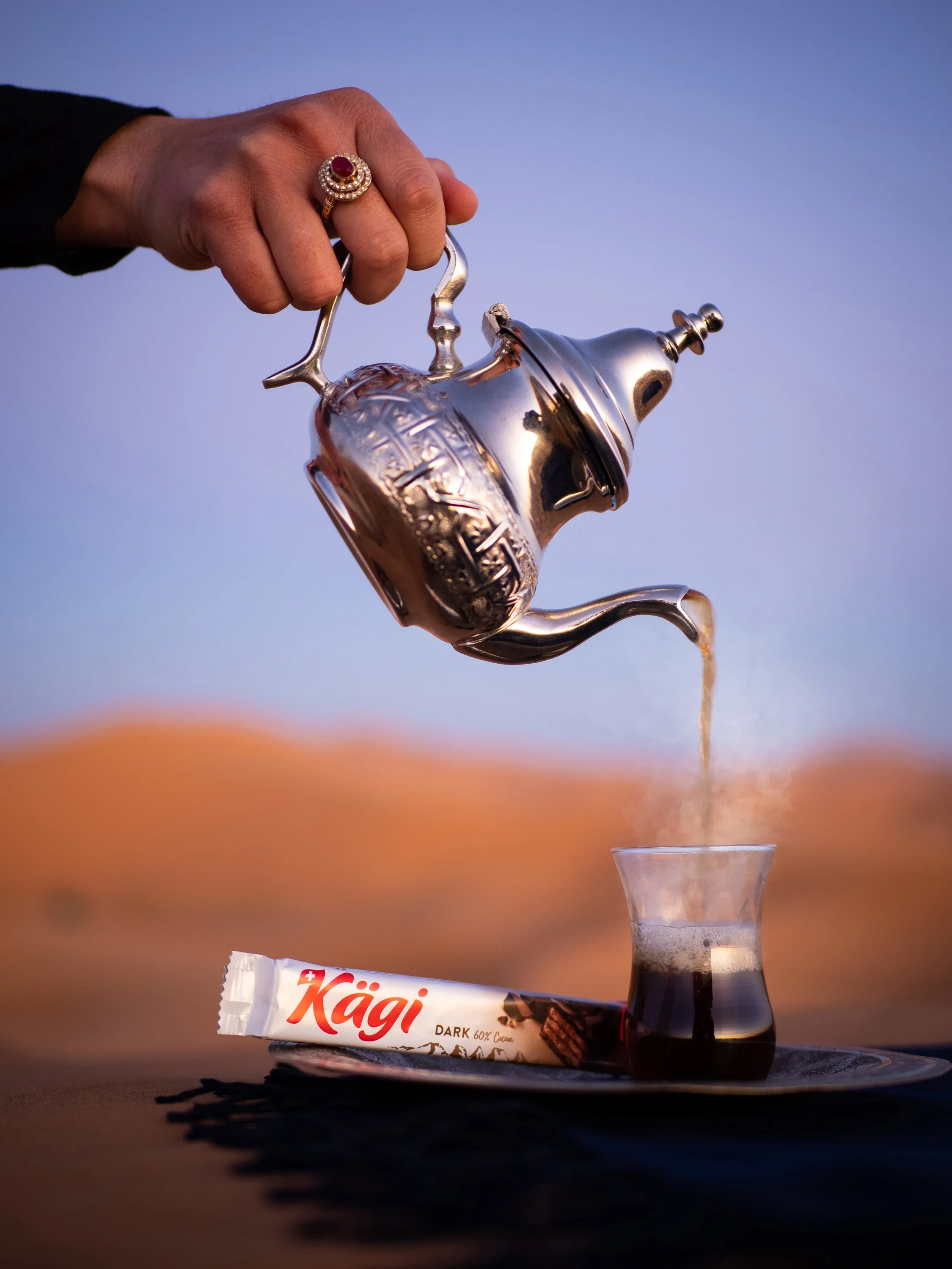 Eine Hand hält eine silberne Kaffeekanne, aus der Kaffee in ein Glas Under demselben fließt. Darunter liegt ein Kagi-Schokoriegel auf einem schwarzen Tablett.
