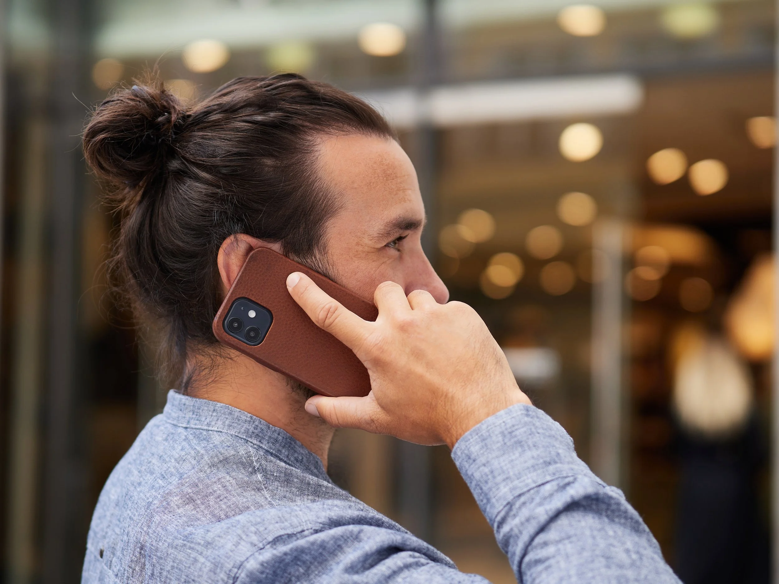 Ein junger Mann mit dunklem, langem Haar in einem Dutt, trägt ein graues Hemd und spricht in ein Smartphone mit brauem Leder-Case, während er im Freien steht.