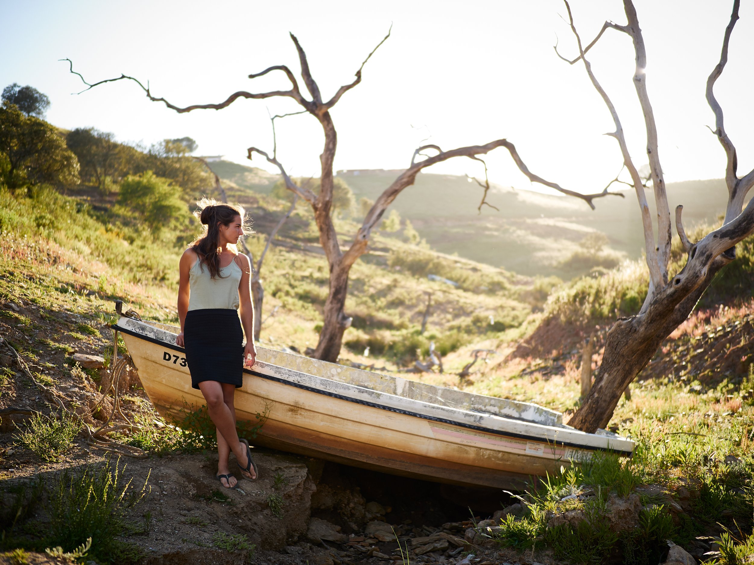 Junge Frau steht an einem alten, verendeten Boot, umgeben von trockenen, windgepeitschten Bäumen in einer ländlichen, sonnigen Landschaft.