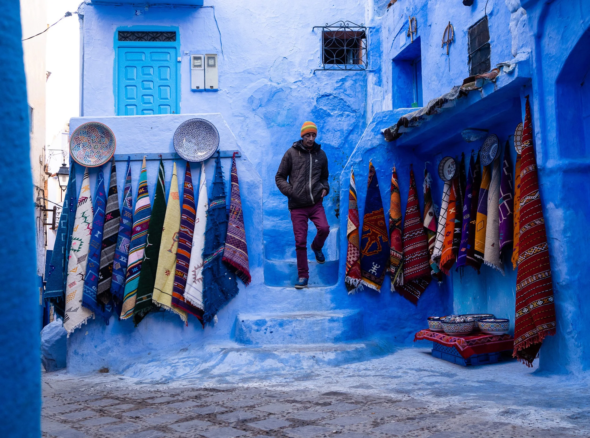 Ein Mann in dunkler Jacke und bunter Mütze läuft durch eine blaue, enge Gasse mit bunten handgearbeiteten Teppichen und Tüchern, die an Wänden aus blauer Farbe hängen, in einer marokkanischen Stadt.