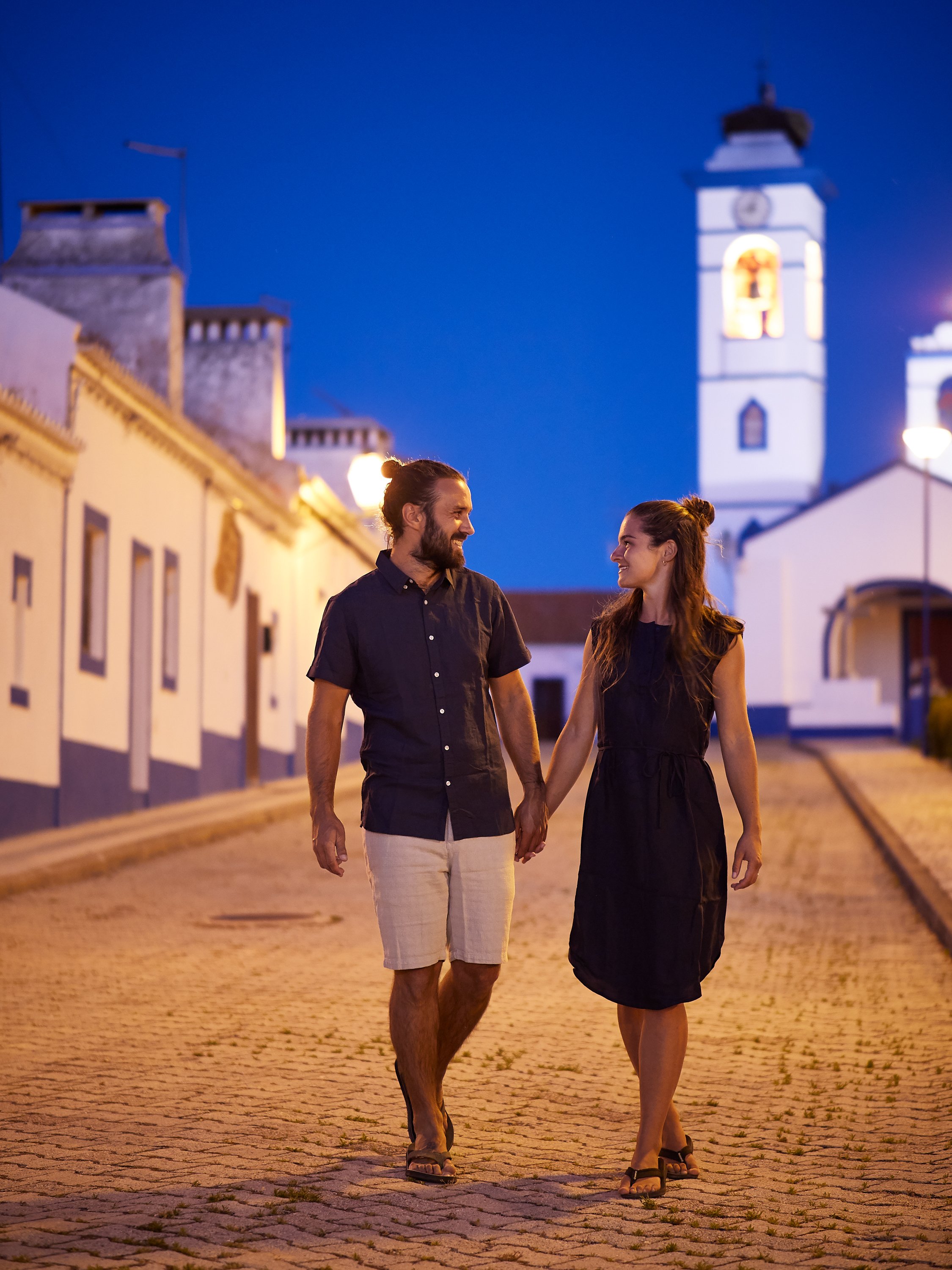 Ein Paar spaziert abends hand-in-Hand durch eine historische Stadt mit weißen Gebäuden und einem Kirchturm im Hintergrund, beleuchtet von Straßenlaternen.
