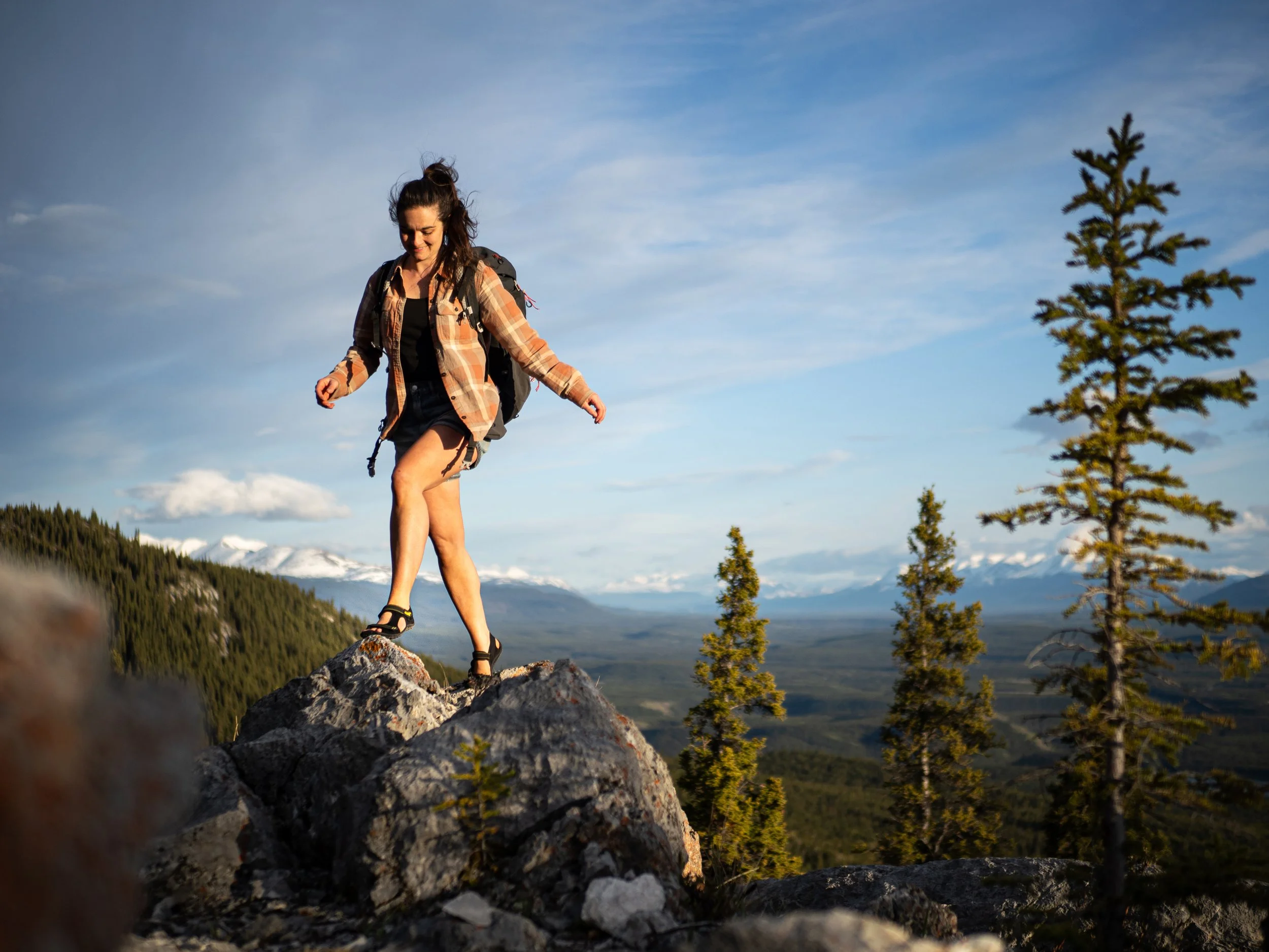 Frau beim Bergwandern auf einem Felsen mit Blick auf eine Landschaft mit Tannenbäumen, Bergen und einer Panorama-Himmel, Sonnenlicht beleuchtet sie. Selbstschutzbekleidung und Rucksack sichtbar.