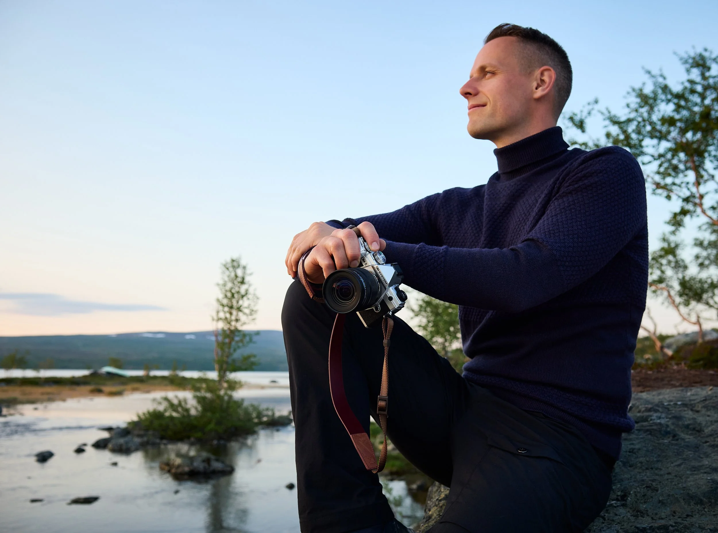 Mann sitzt am Flussufer mit Kamera in der Hand, Blick in die Ferne, bei Sonnenuntergang, ruhige Naturumgebung mit Wasser, Steinen und Bäumen