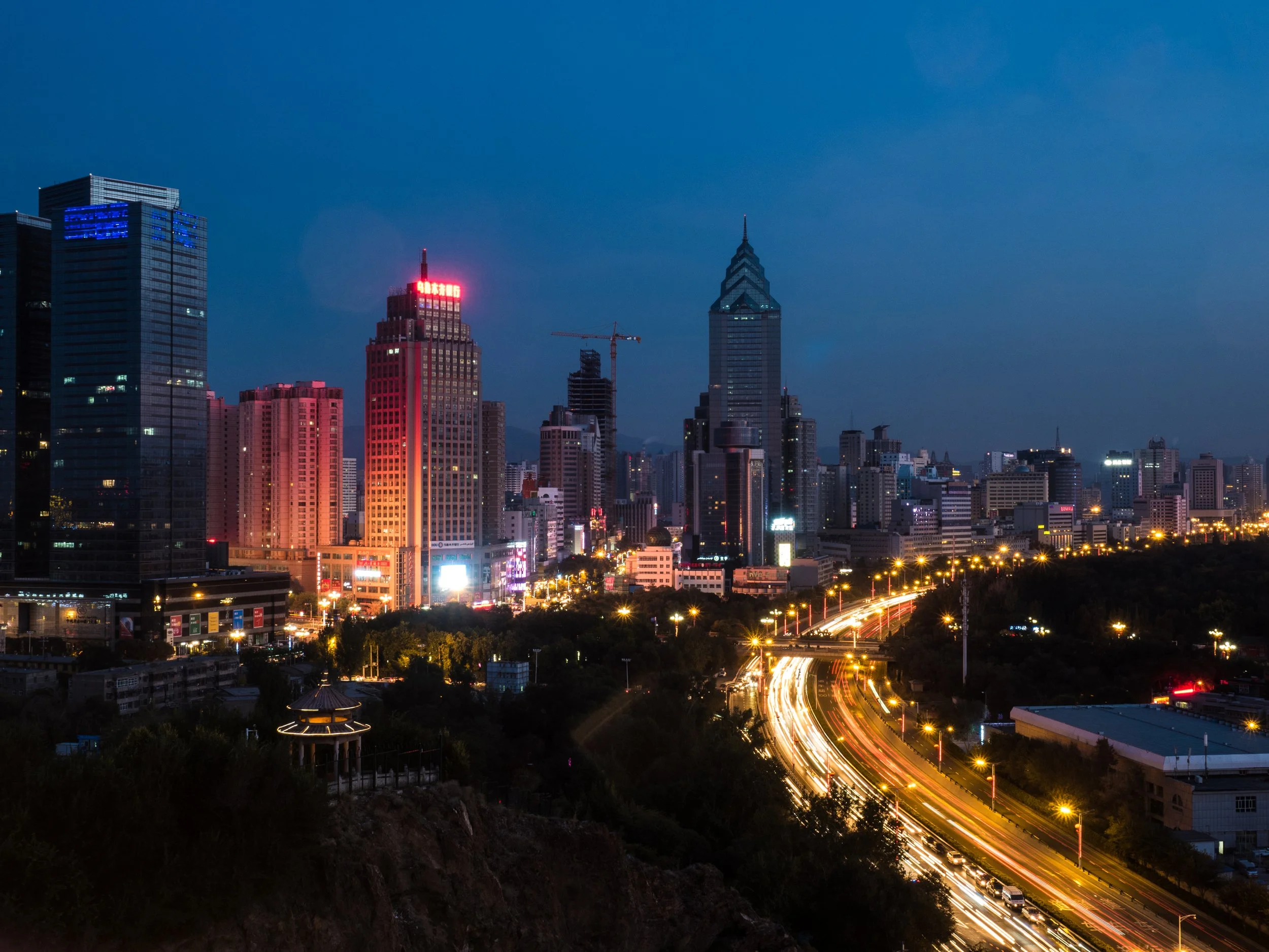 Abendliche Skyline einer Stadt mit leuchtenden Hochhäusern, beleuchteter Autobahn mit Lichtspuren, und dunklem Himmel.