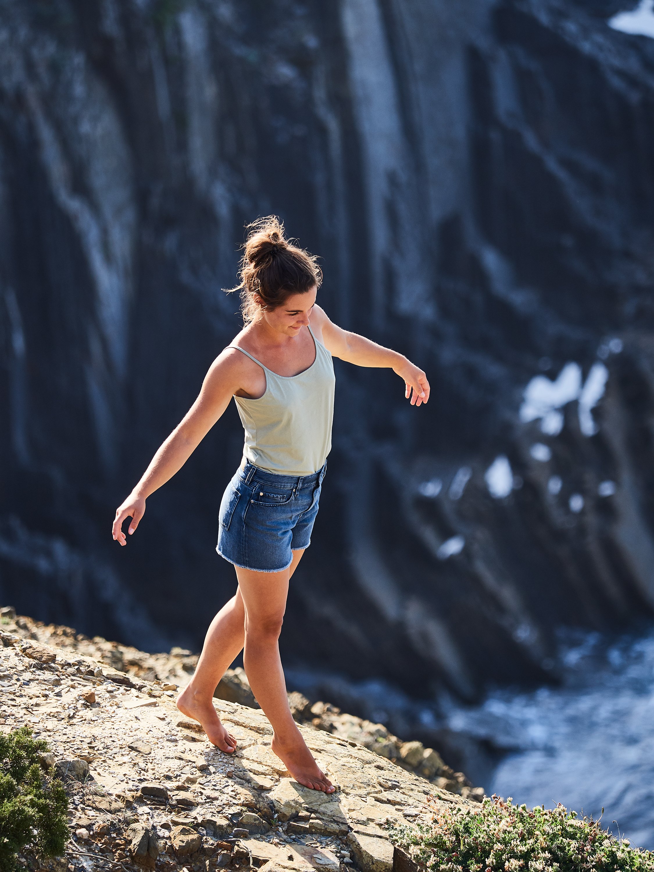 Eine junge Frau in einem weißen Tanktop und Jeansshorts balanciert barfuß auf einem schmalen, felsigen Pfad neben einem Wasserfall.