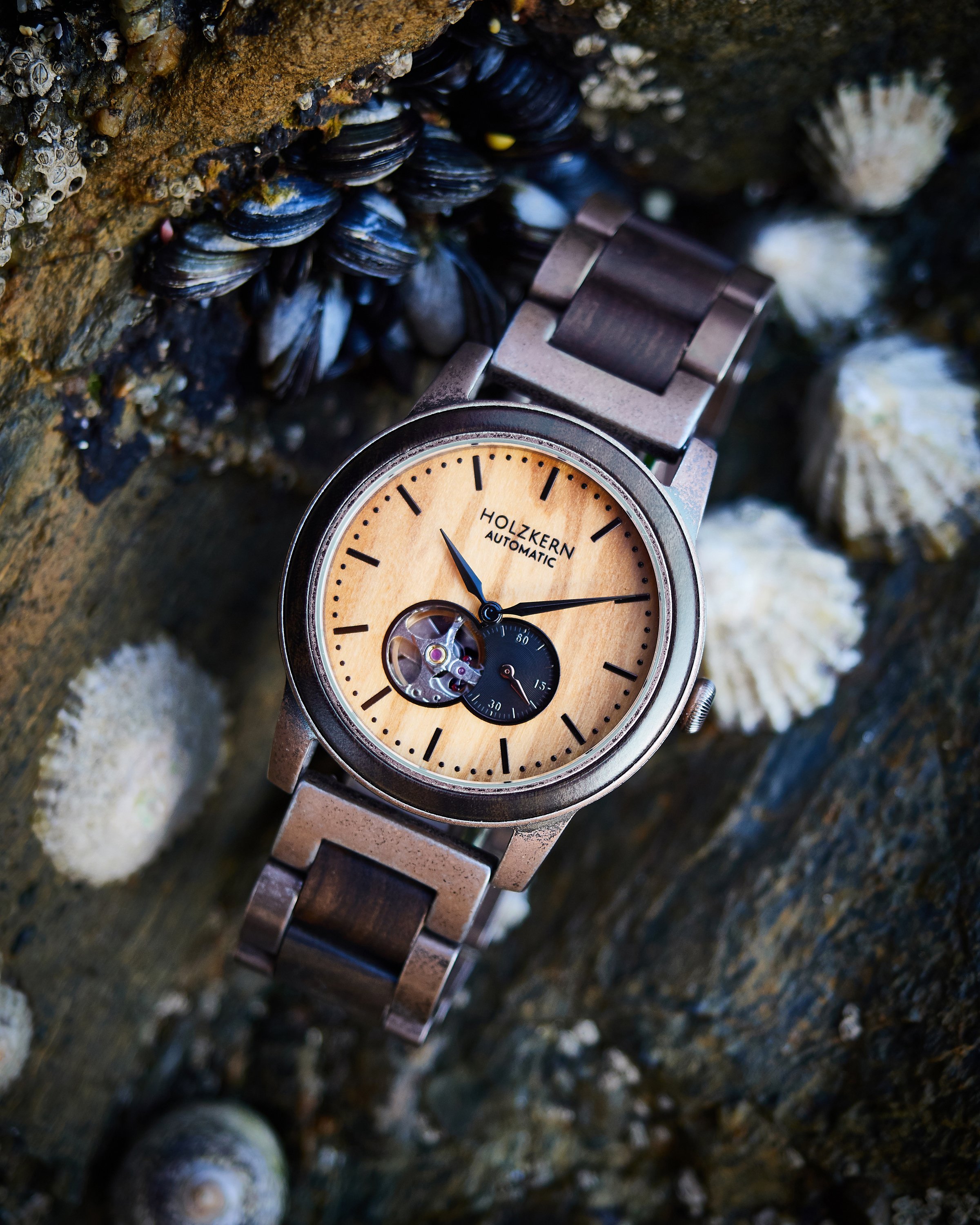 Eine Holzkern-Automatikuhr mit silbernem Armband, auf einer felsigen Oberfläche mit Muscheln. Das Zifferblatt ist beige mit schwarzen Markierungen und Zeigern, und es gibt ein Sichtfenster des Uhrwerks sowie eine kleine Sekunde-Anzeige.