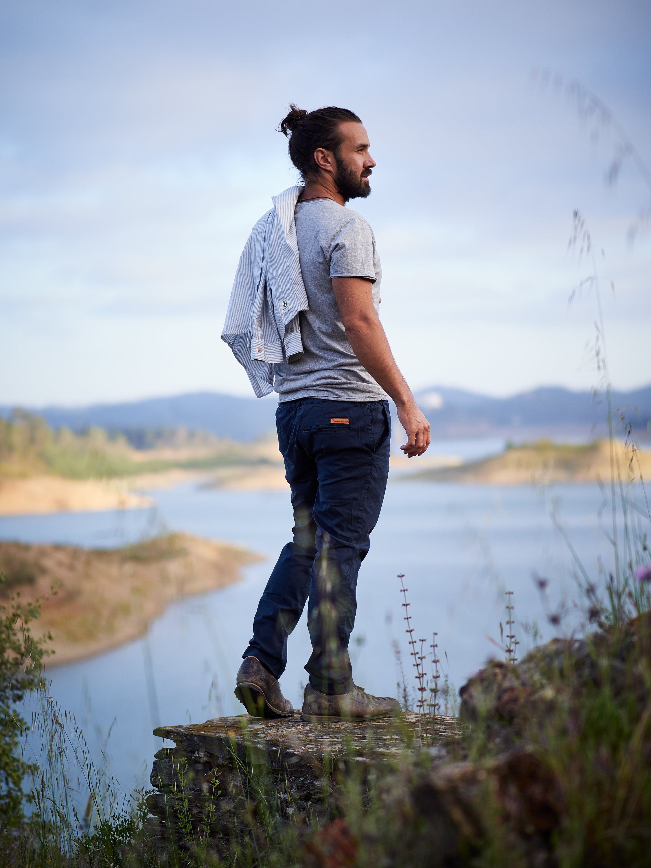 Ein Mann mit dunklem Haar und Bart, der auf einem Felsen neben einem Fluss oder See steht, während er die Aussicht genießt.