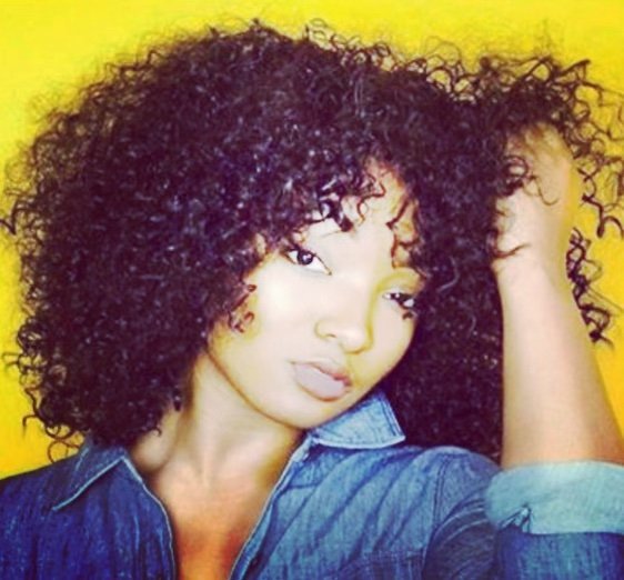 A woman with voluminous, curly black hair taking a selfie against a yellow background.