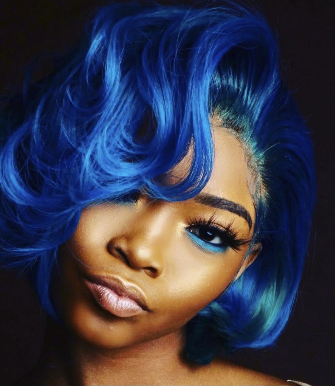 Close-up of a woman with vibrant blue hair, bold makeup, and a confident expression.