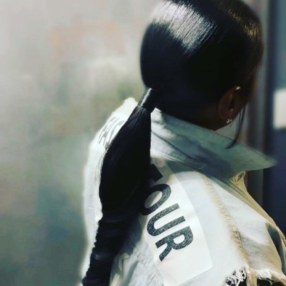 A woman with long, straight black hair tied in a low ponytail, wearing a light-colored jacket with a patch that says 'YOUR'. She is standing sideways near a window or mirror, with her head turned away from the camera.