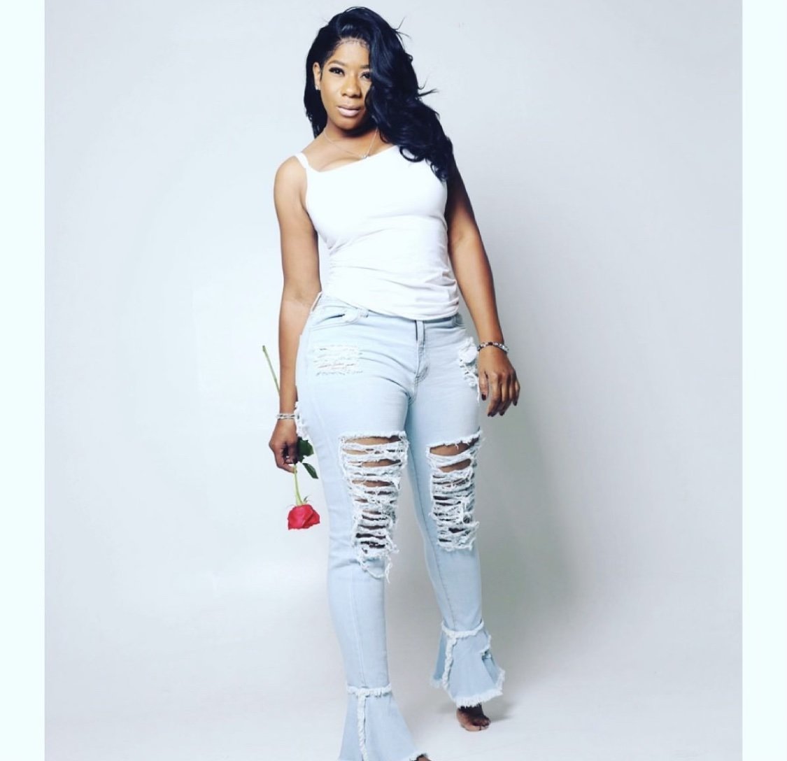 A woman with long black hair stands against a plain light-colored background, holding a red rose in her right hand, wearing a white tank top and heavily distressed light blue jeans.