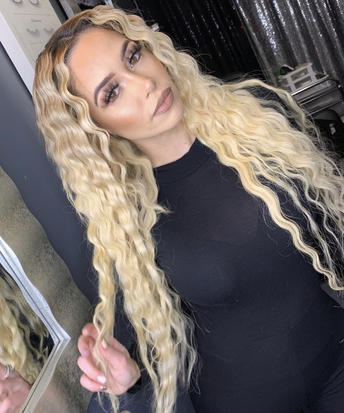 woman with long, wavy blonde hair, wearing a black top, posing in front of a mirror in a salon or makeup studio.