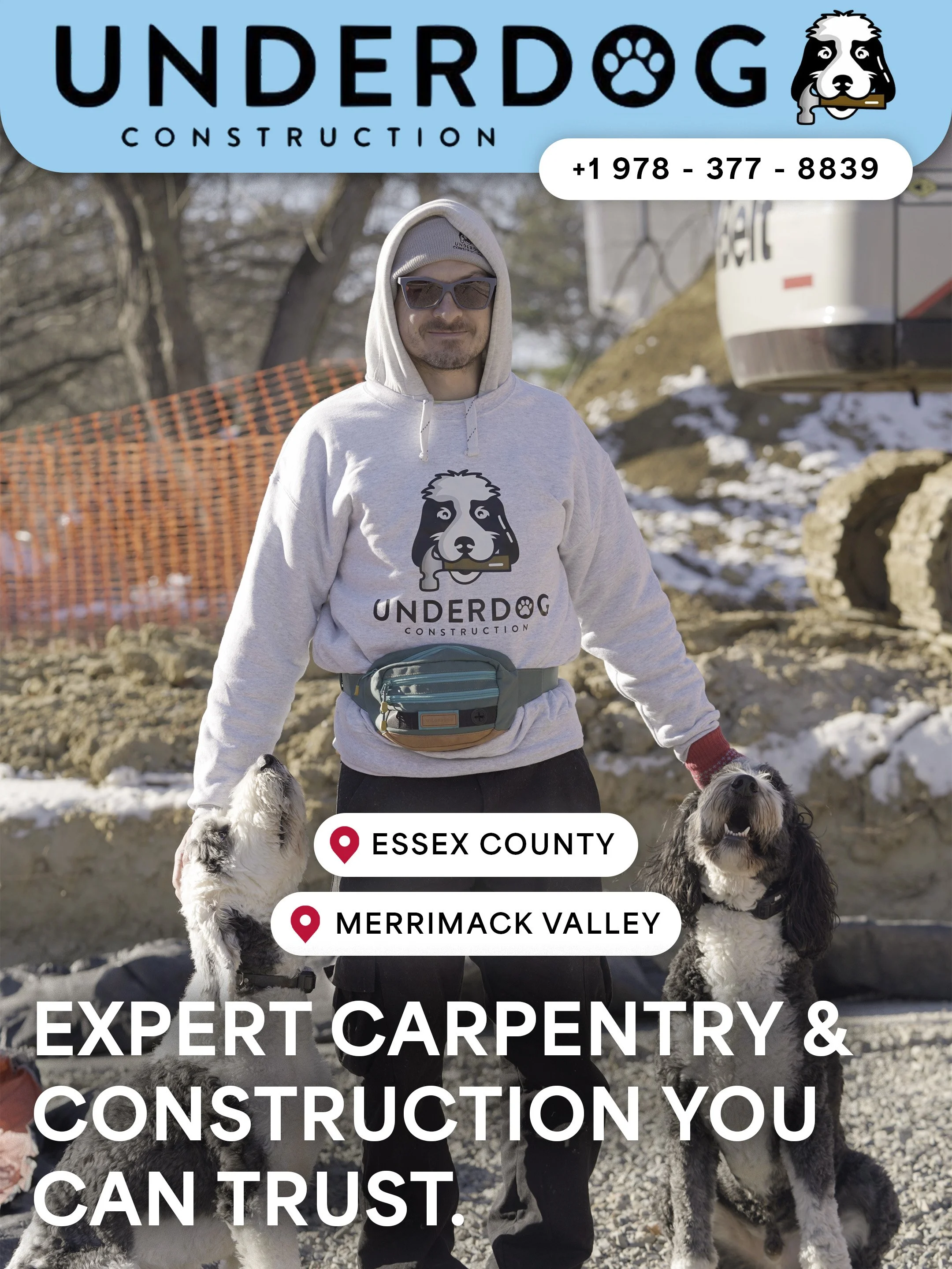 A man wearing sunglasses and a hoodie with the UnderDog Construction logo, standing outdoors with three dogs on a construction site with snow and rocks.