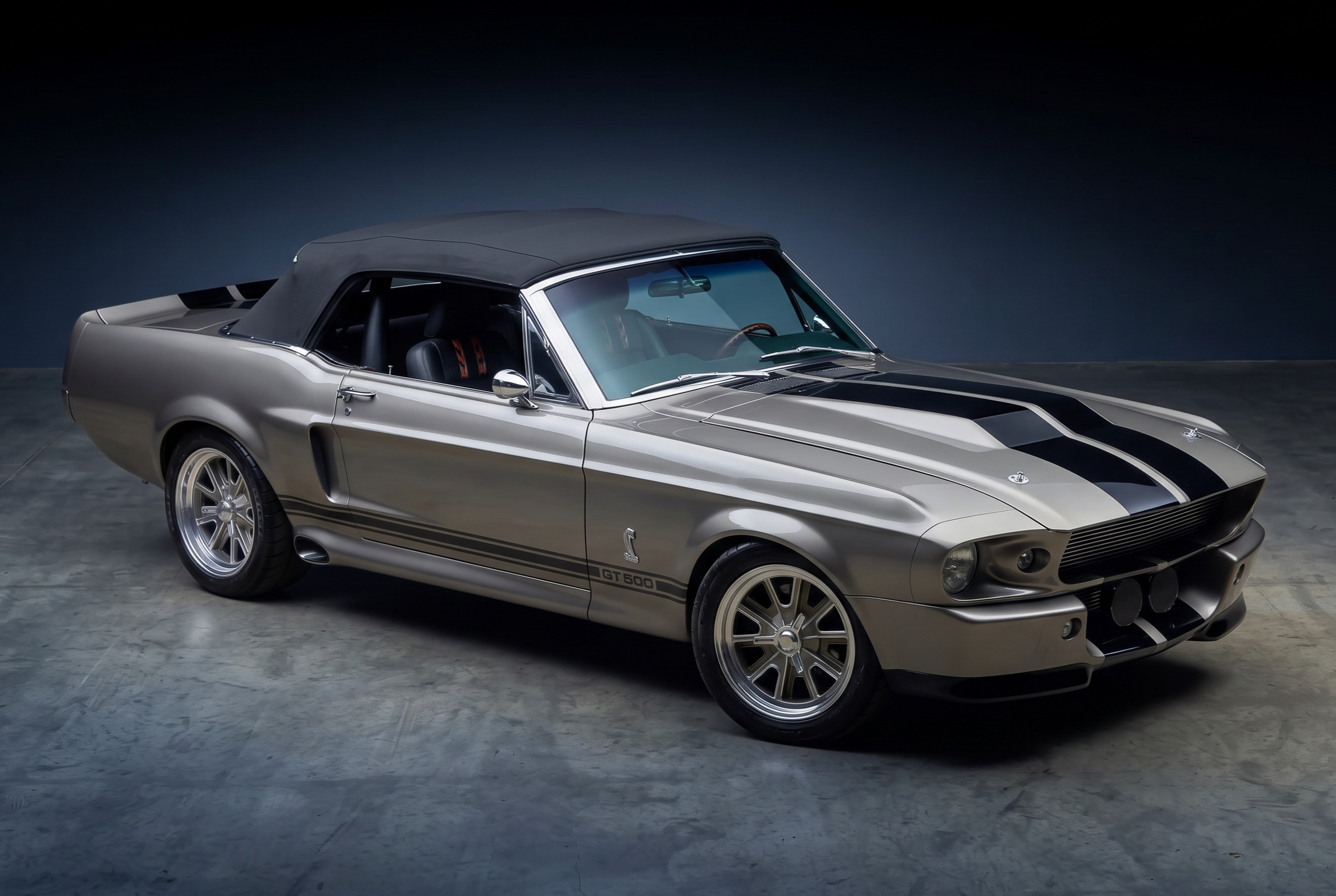 A vintage silver Ford Mustang Shelby GT500 with black racing stripes, parked on a gray concrete floor against a dark background.