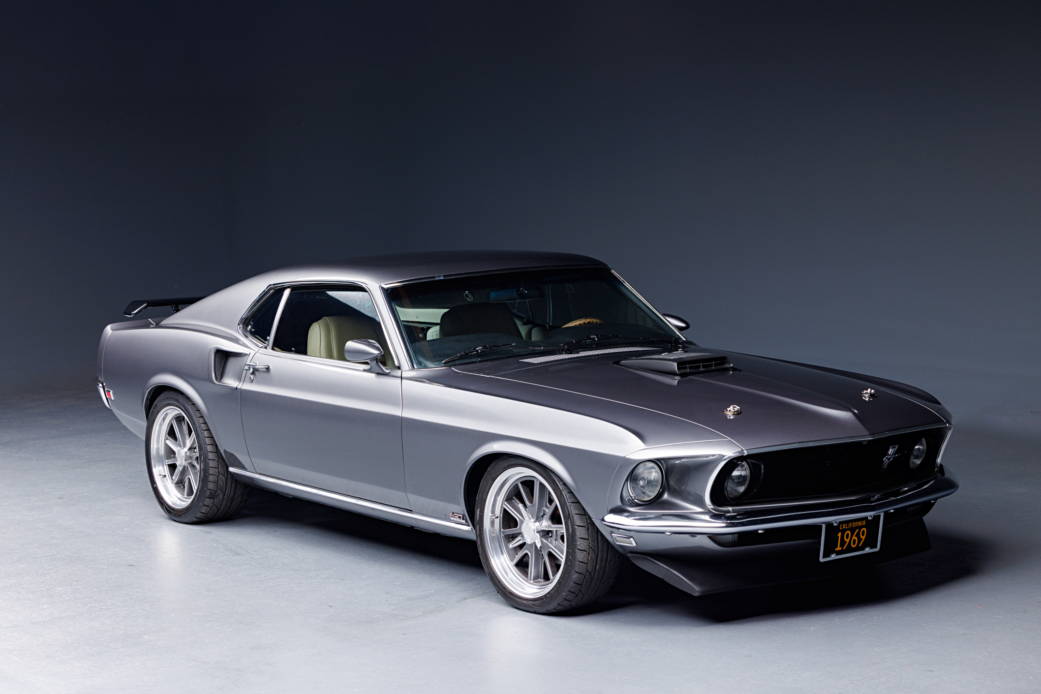 A silver 1969 Ford Mustang coupe with black racing stripes, parked indoors against a dark background.
