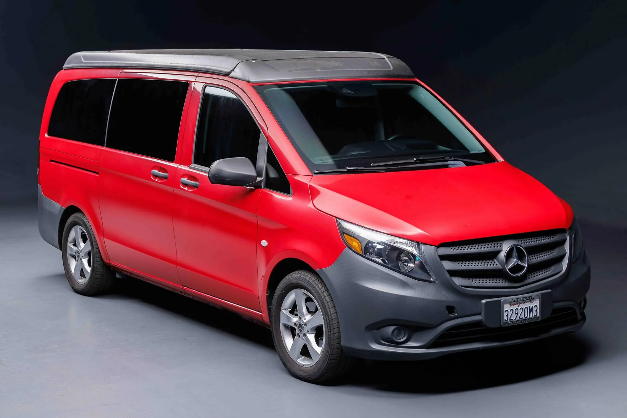 Red Mercedes-Benz van with a gray roof and tinted windows, parked on a gray surface against a dark background.
