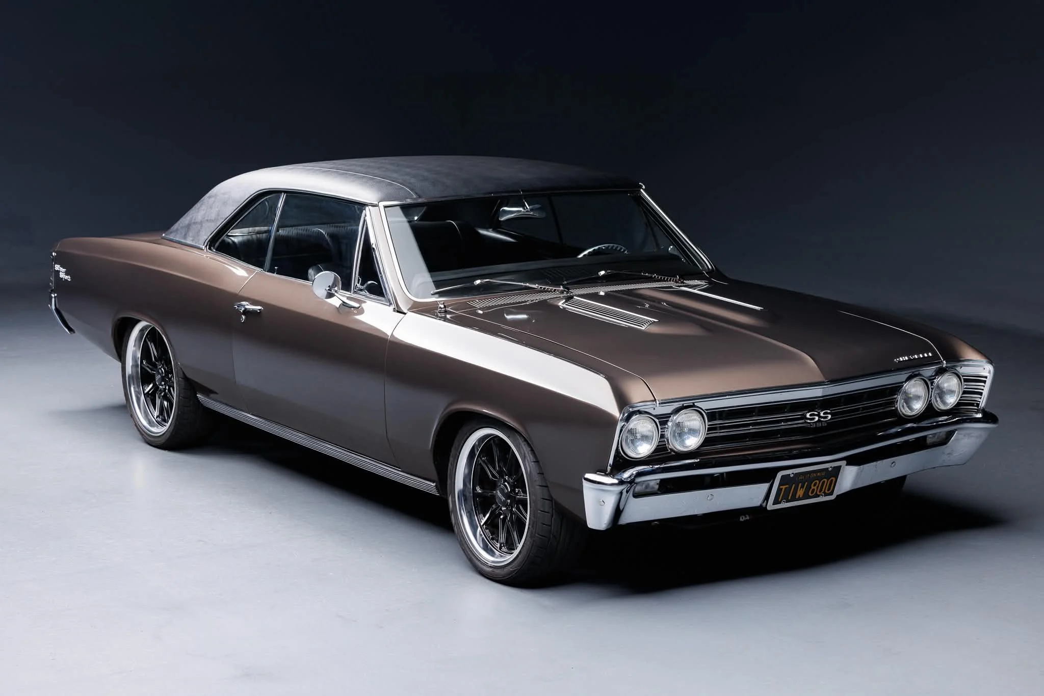 A vintage Chevrolet SS 396 car with a two-tone paint job, featuring a brown body and a silver roof, parked in a studio with a dark background.