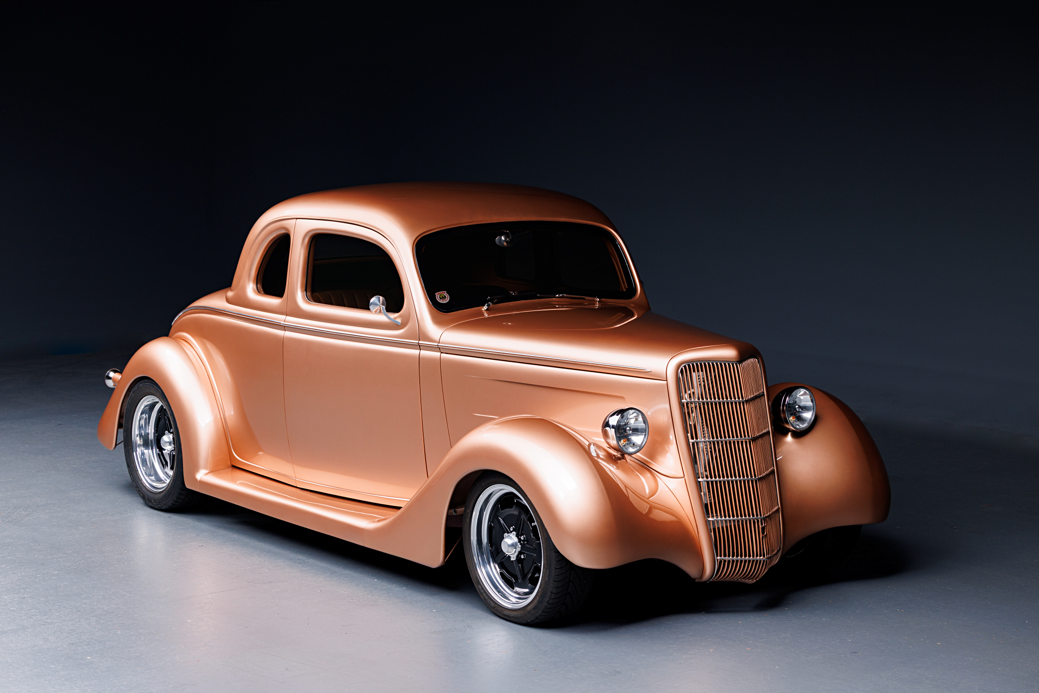 A vintage-style car with a shiny metallic copper finish, rounded body, large front grille, and covered rear wheels, positioned in a studio against a dark background.