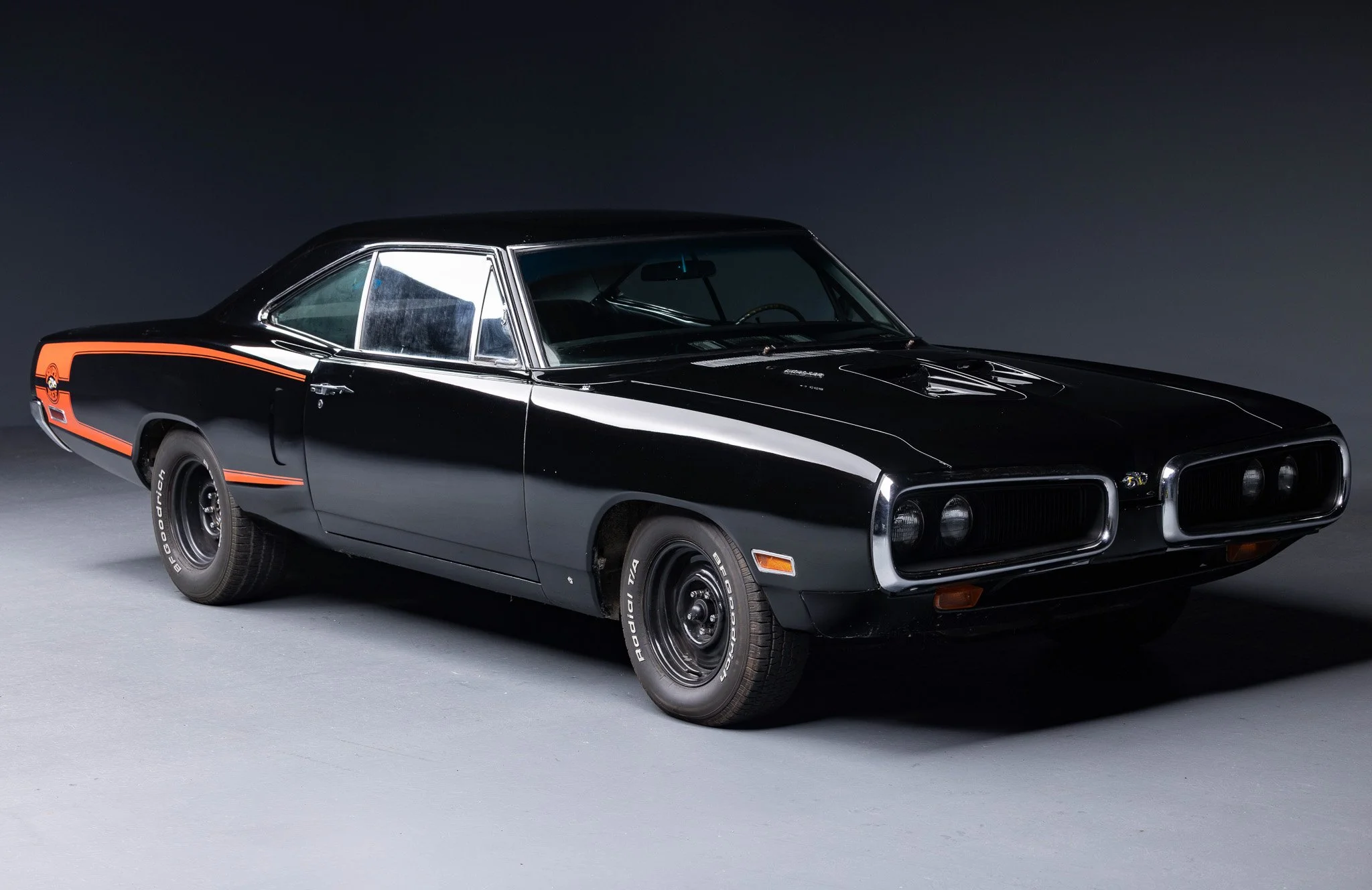 A vintage black muscle car with orange stripe and Chevy logo on the side, parked on a gray surface against a dark background.