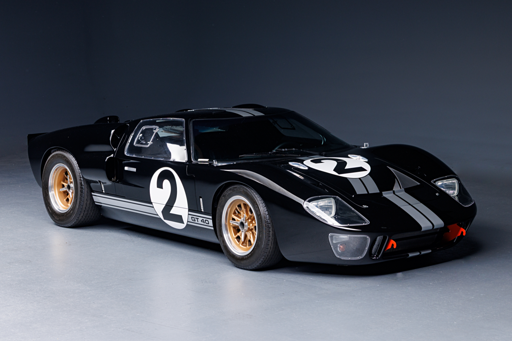 A classic black racing car with the number 2 on its hood and sides, gold wheels, and white racing stripes, displayed on a smooth gray surface.