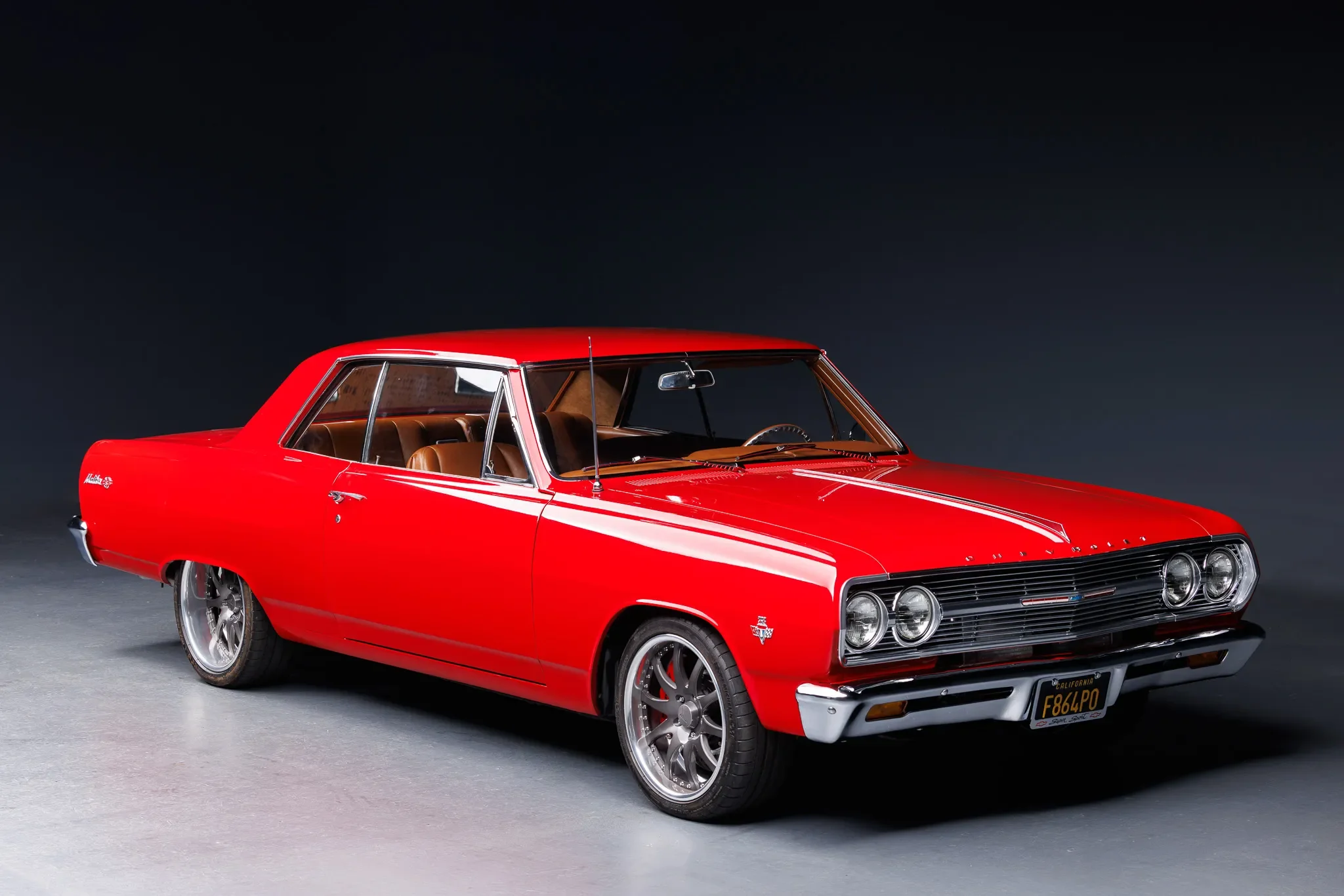 A vintage red muscle car with tan interior, chrome rims, and a California license plate, set against a dark background.