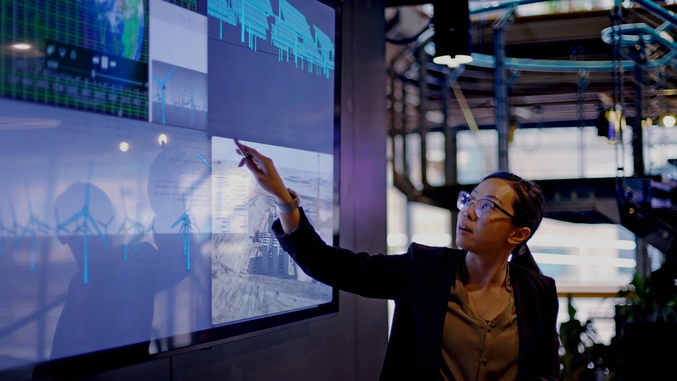 Woman pointing at large presentation screen with graphs and wind turbines, in a modern indoor setting with industrial design elements.