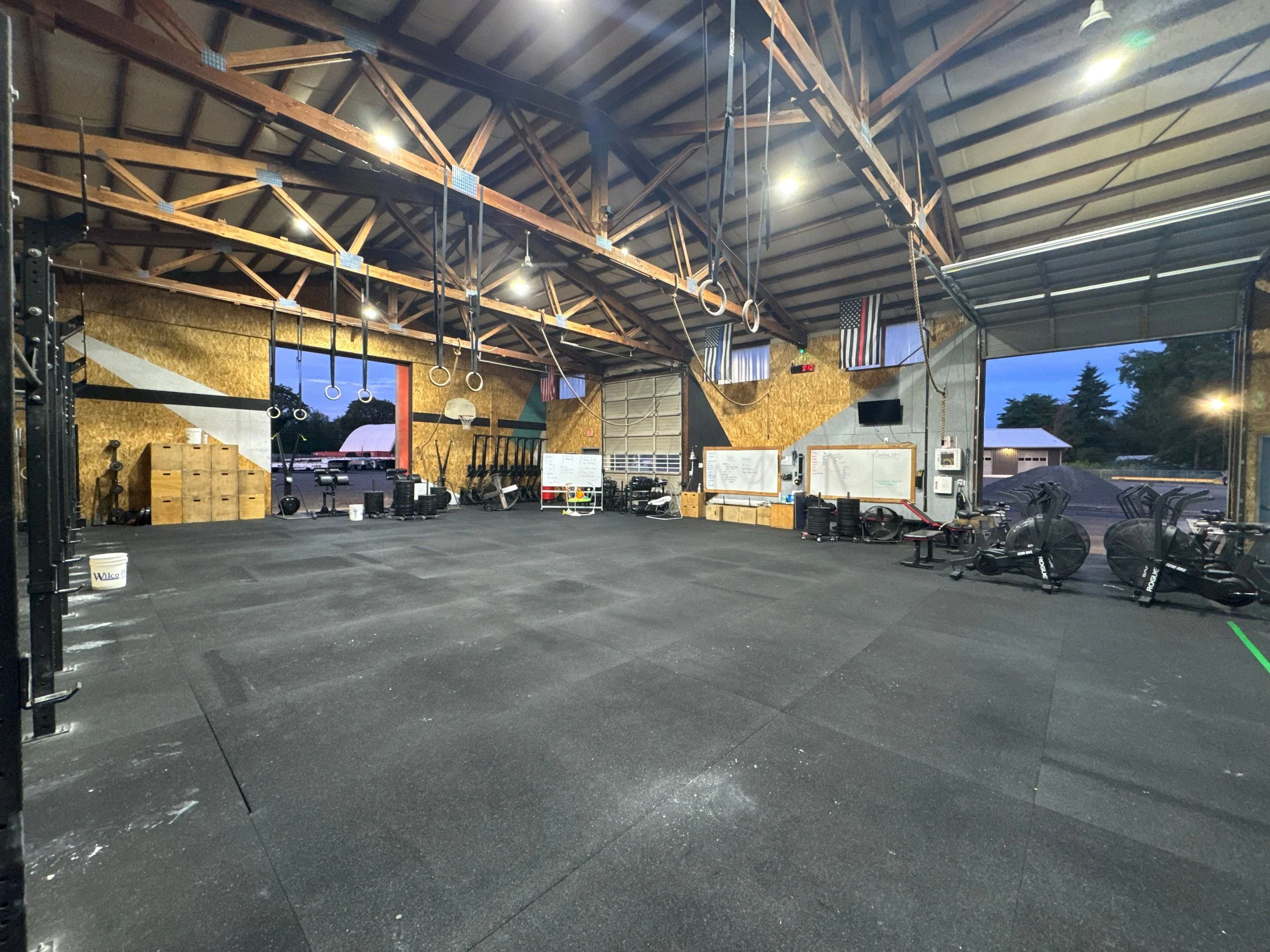 Empty CrossFit gym with black rubber flooring, gym equipment, open garage door, and American flags on the wall