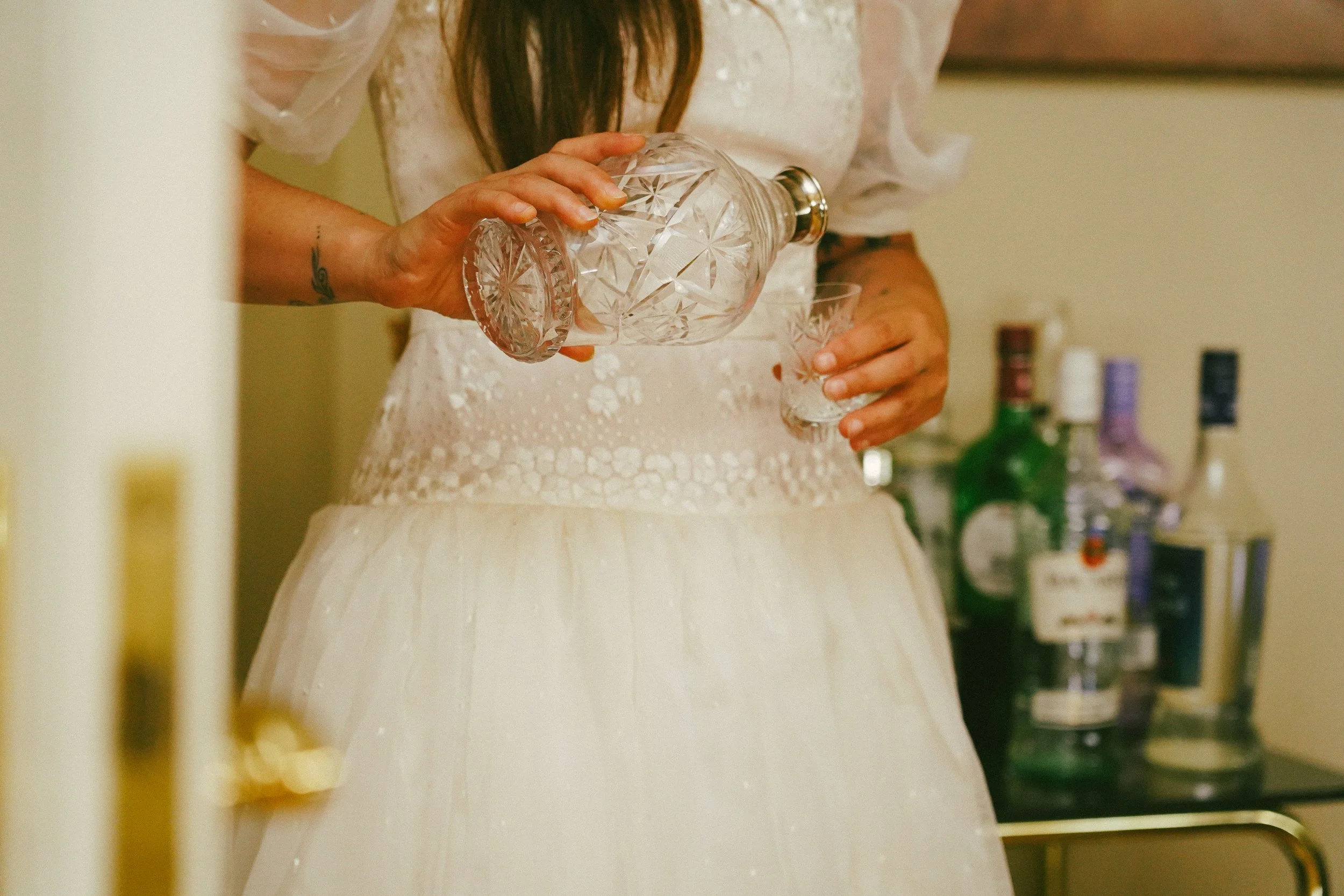 Una mujer en vestido blanco sirviendo alcohol en un vaso usando una botella de cristal cortado.