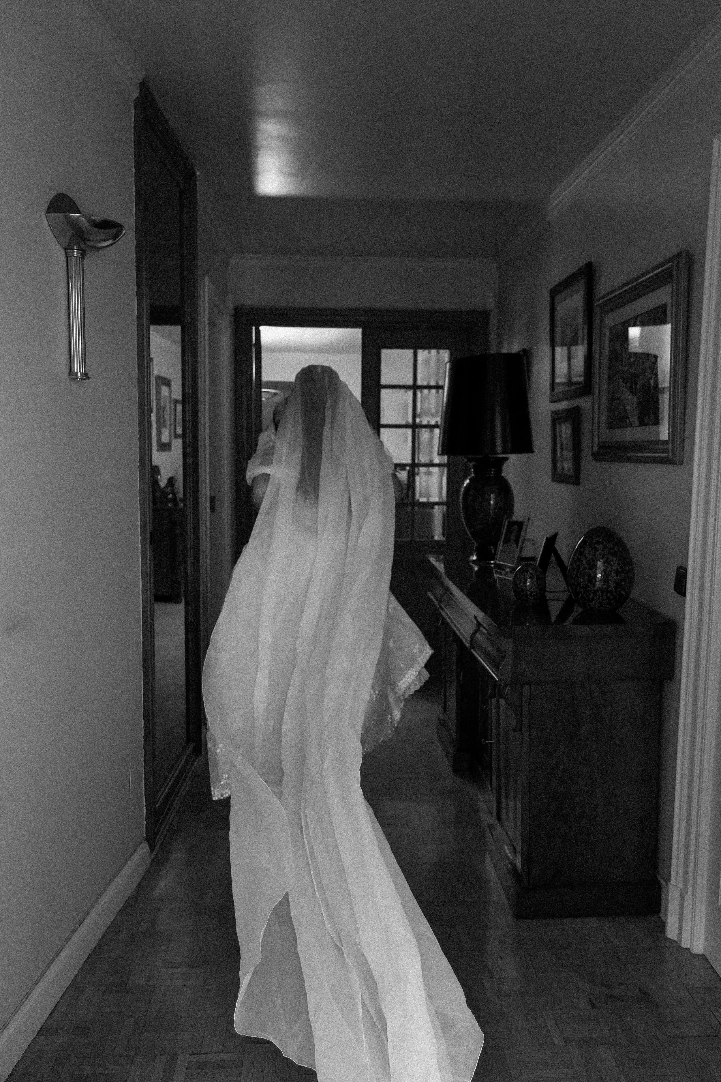 Una figura con un vestido y velo de novia de pie en un pasillo en blanco y negro, frente a una puerta con ventanas.
