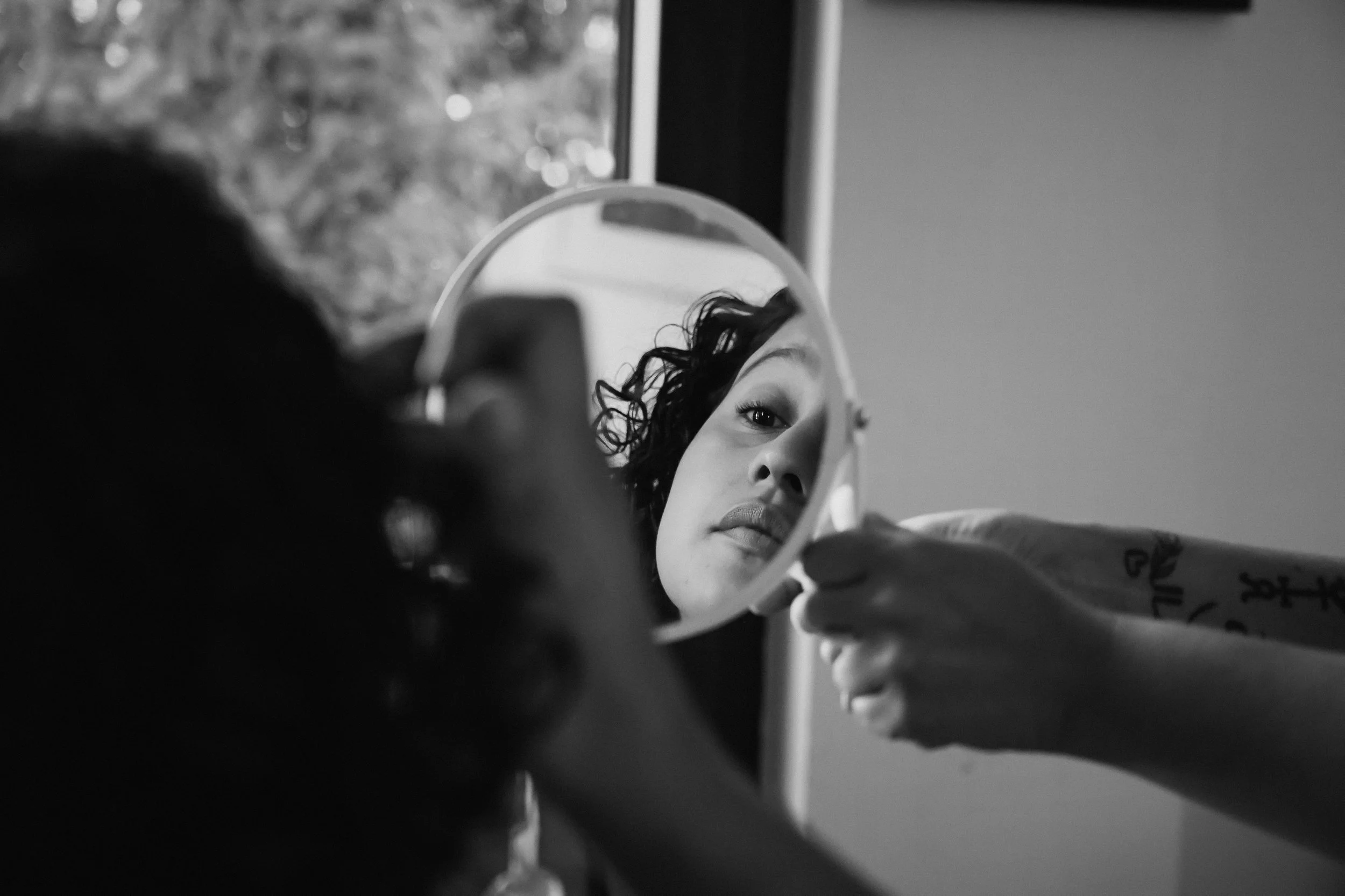 Una mujer mirando su reflejo en un espejo circular, sosteniendo el espejo con una mano, con cabello rizado y expresión facial neutra, en un entorno interior.