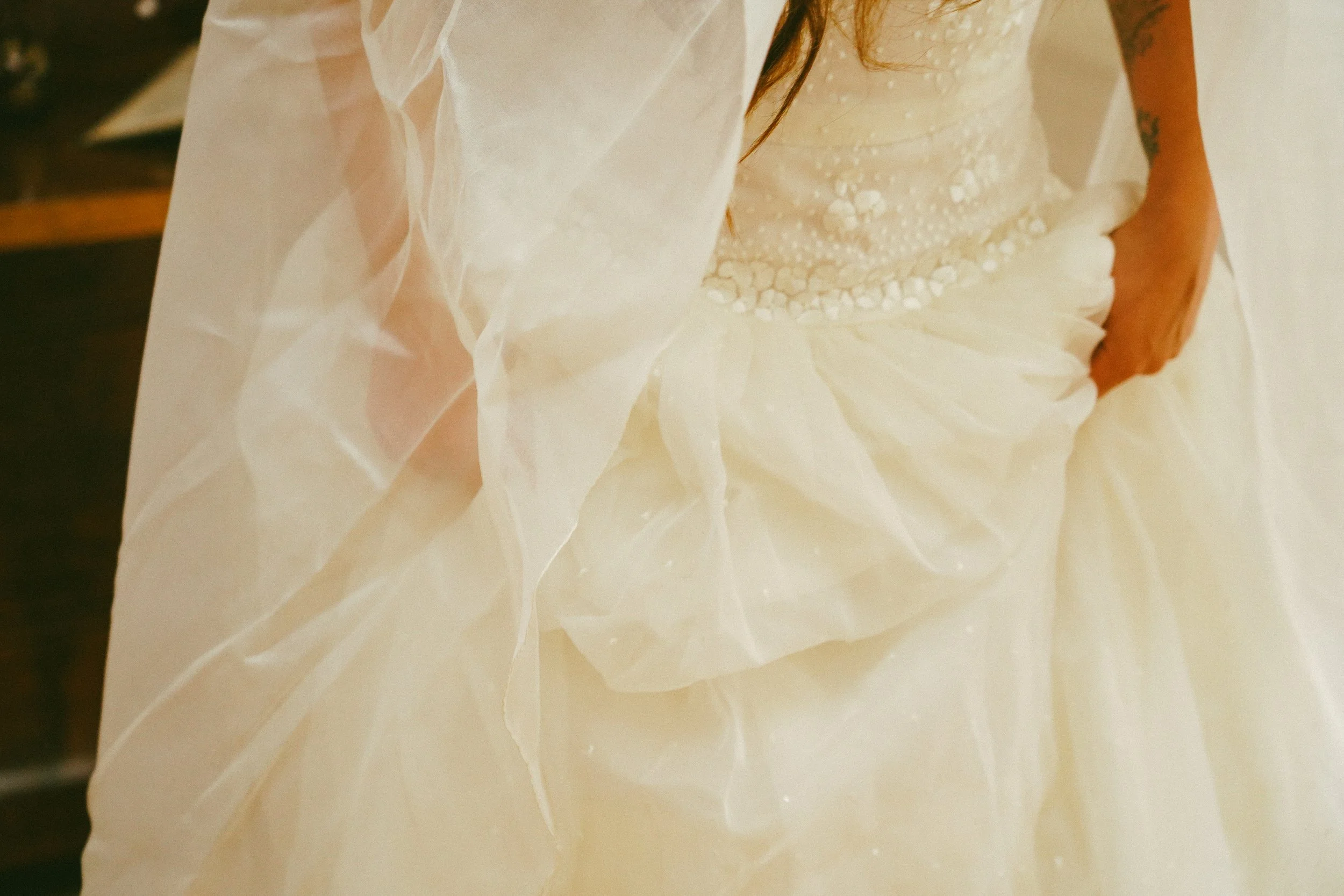 Vestido de novia con encaje y volantes en la falda, sosteniendo la falda con una mano, en un entorno interior.