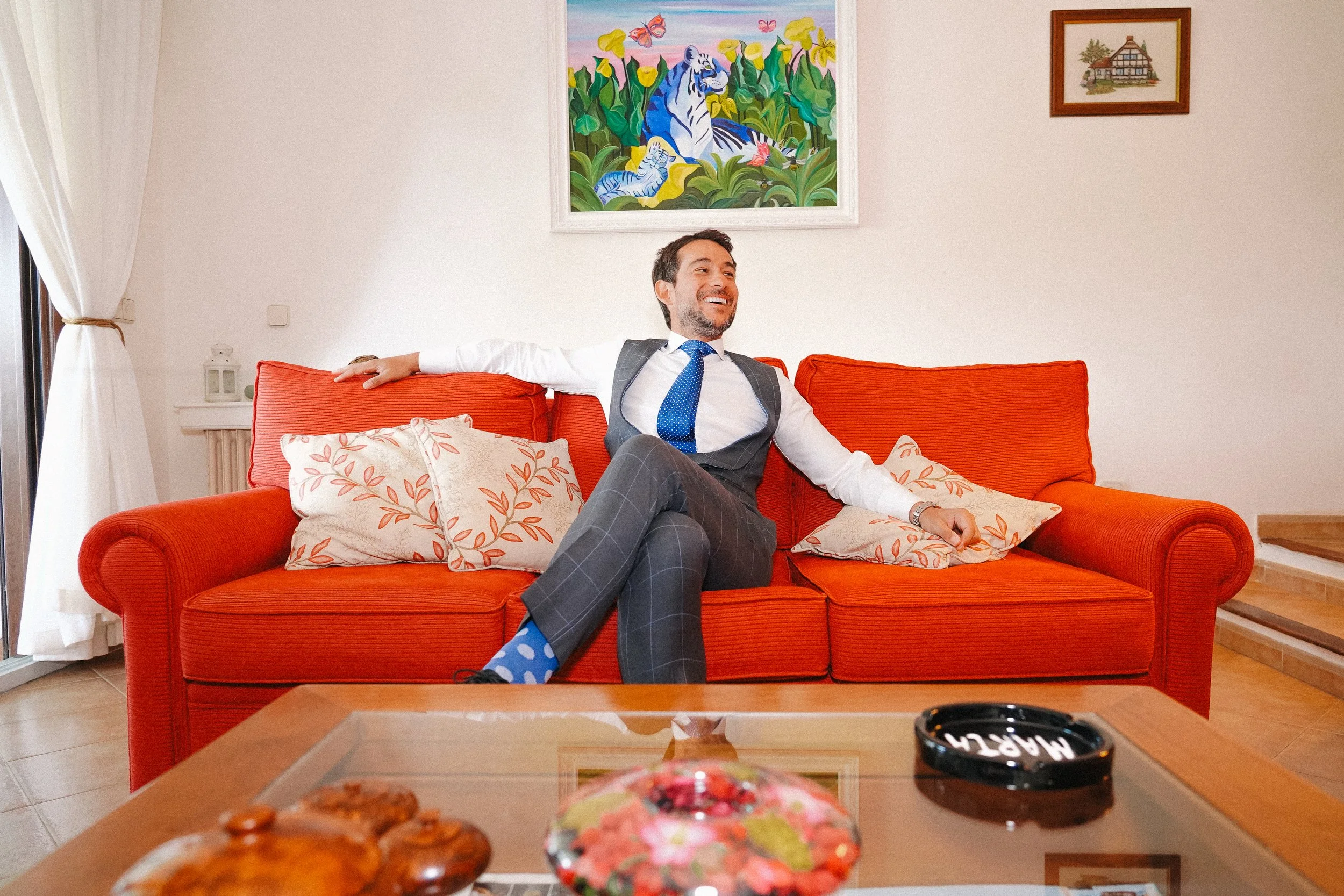 Hombre sonriendo sentado en un sofá naranja en una sala de estar, usando un traje de negocios y calcetas de puntos azules, con cuadros coloridos en la pared y una mesa de centro con objetos decorativos y una cenicero.